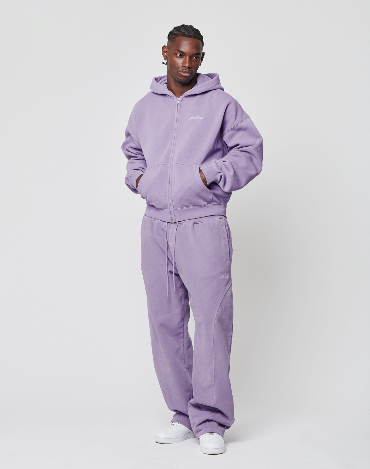 A man wears the LIVE FAST DIE YOUNG Basic Script Hooded Zip and matching sweatpants in light purple with Vintage-Waschung and subtle logo-stick details, standing against a plain white background with his hands in his pockets.