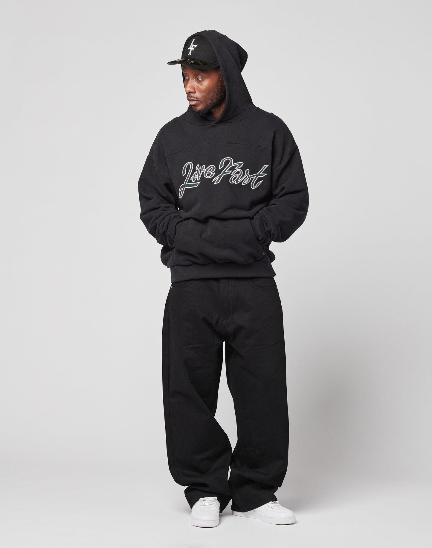 A man wears the LFDY Script Hooded sweatshirt by LIVE FAST DIE YOUNG with an oversize fit and distress effects, paired with black pants, a black cap, and white sneakers, standing against a plain white background with hands in pockets.