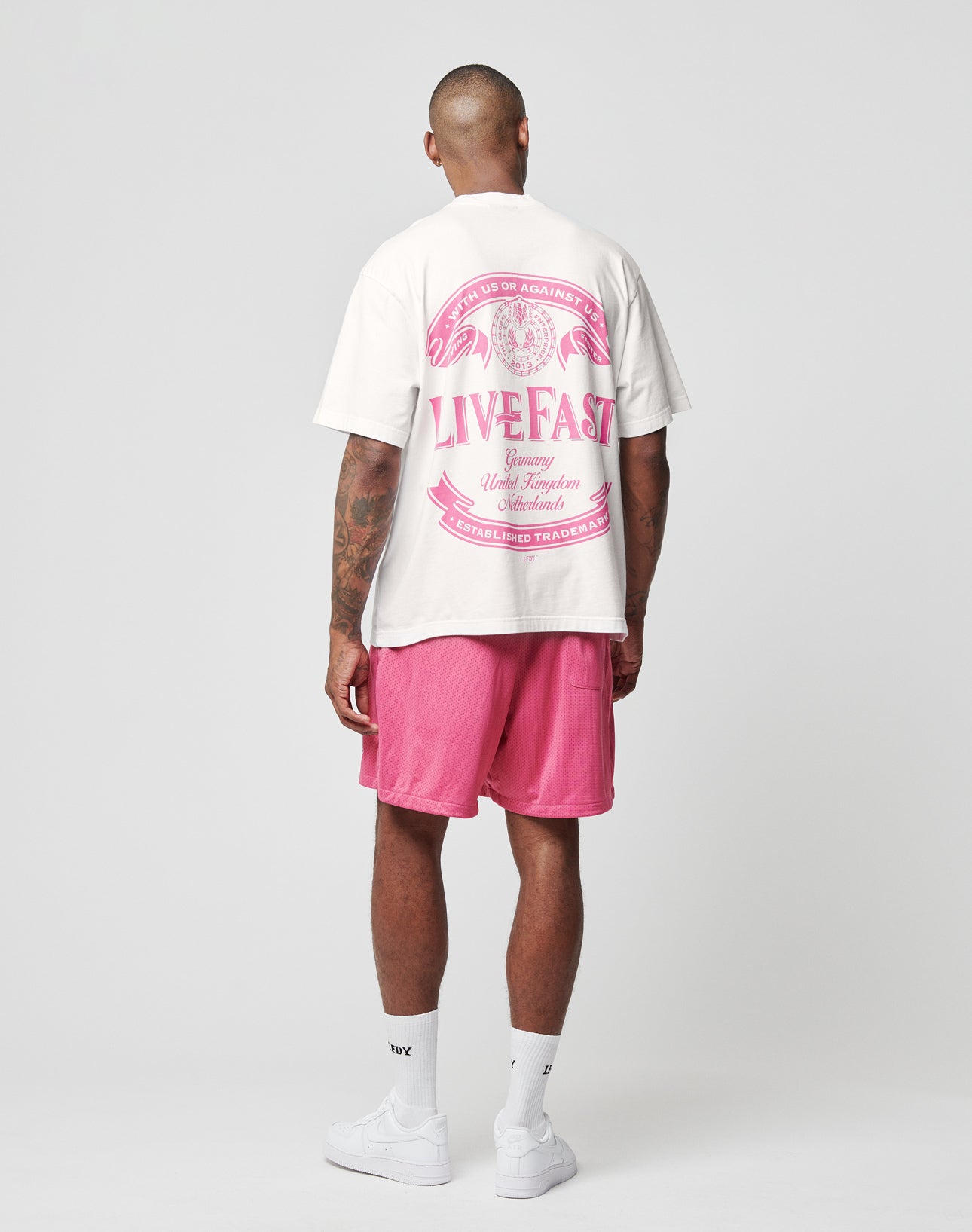 A man faces away, wearing the LFDY Loyal Tee by LIVE FAST DIE YOUNG—an oversized white t-shirt with pink front and back graphics reading LIVE FAST—paired with pink shorts, white socks, and sneakers, set against a light gray background.