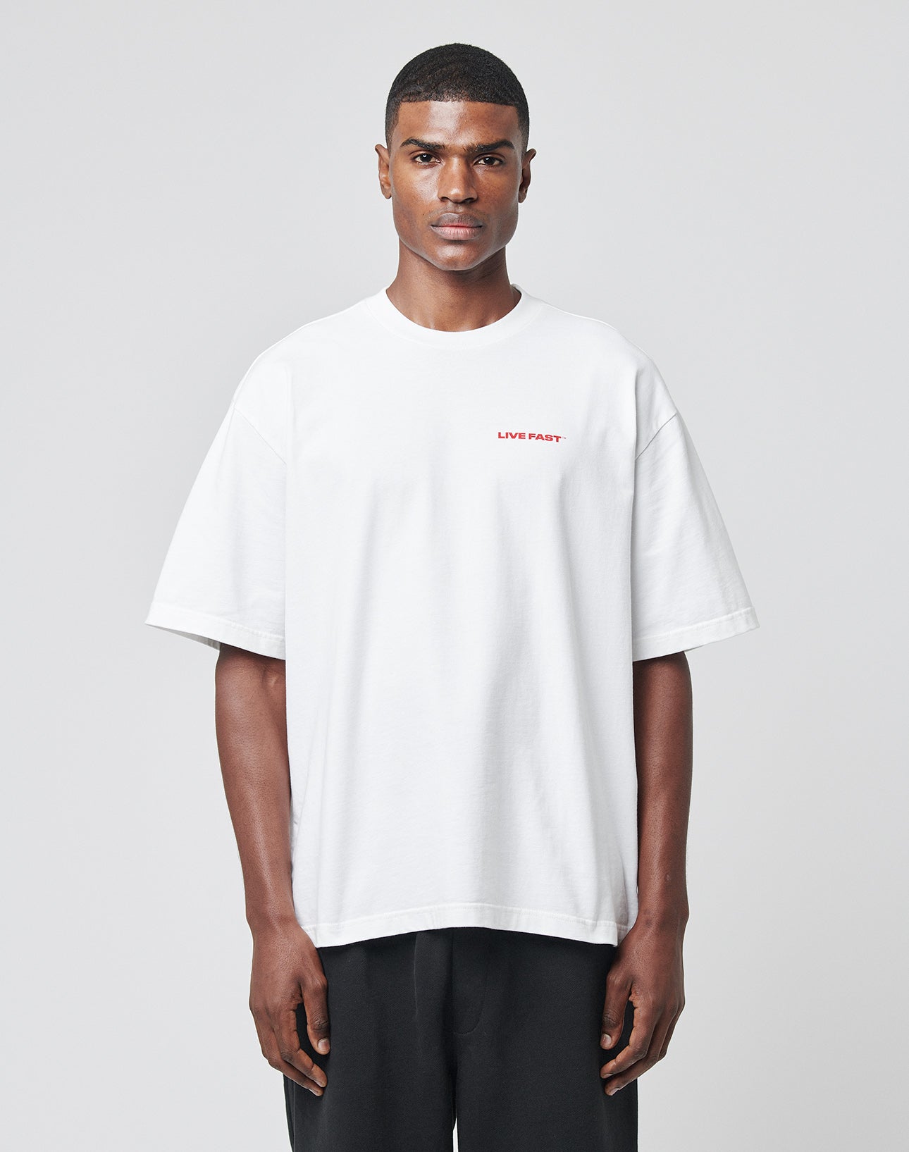 A person faces forward wearing the LFDY Cinema Tee by LIVE FAST DIE YOUNG—a plain white oversized T-shirt with small red LIVE FAST lettering on the left chest, made from 100% cotton—paired with loose black pants on a light gray background.