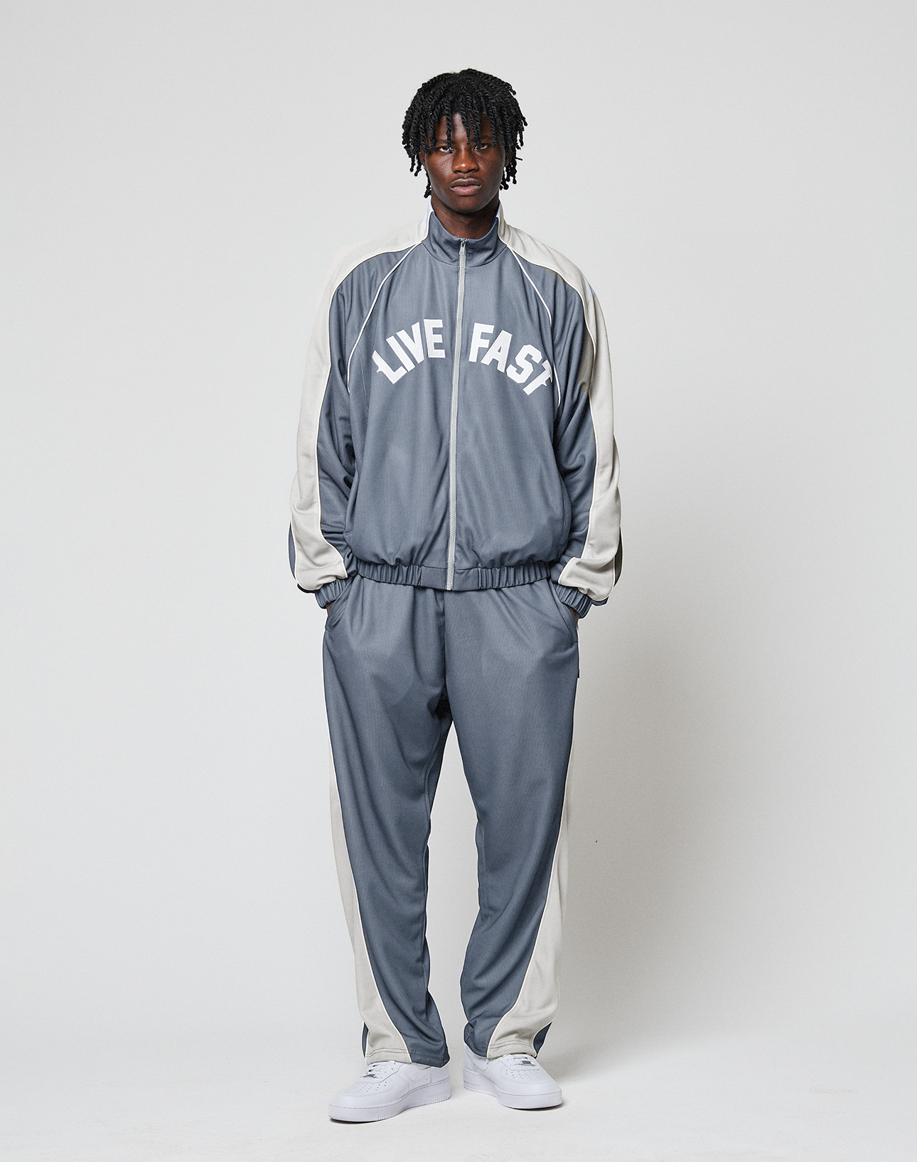 A person stands against a plain background wearing LIVE FAST DIE YOUNG’s gray and white LFDY DY Trackpants with a comfortable elastikbund and wide fit, paired with white sneakers.