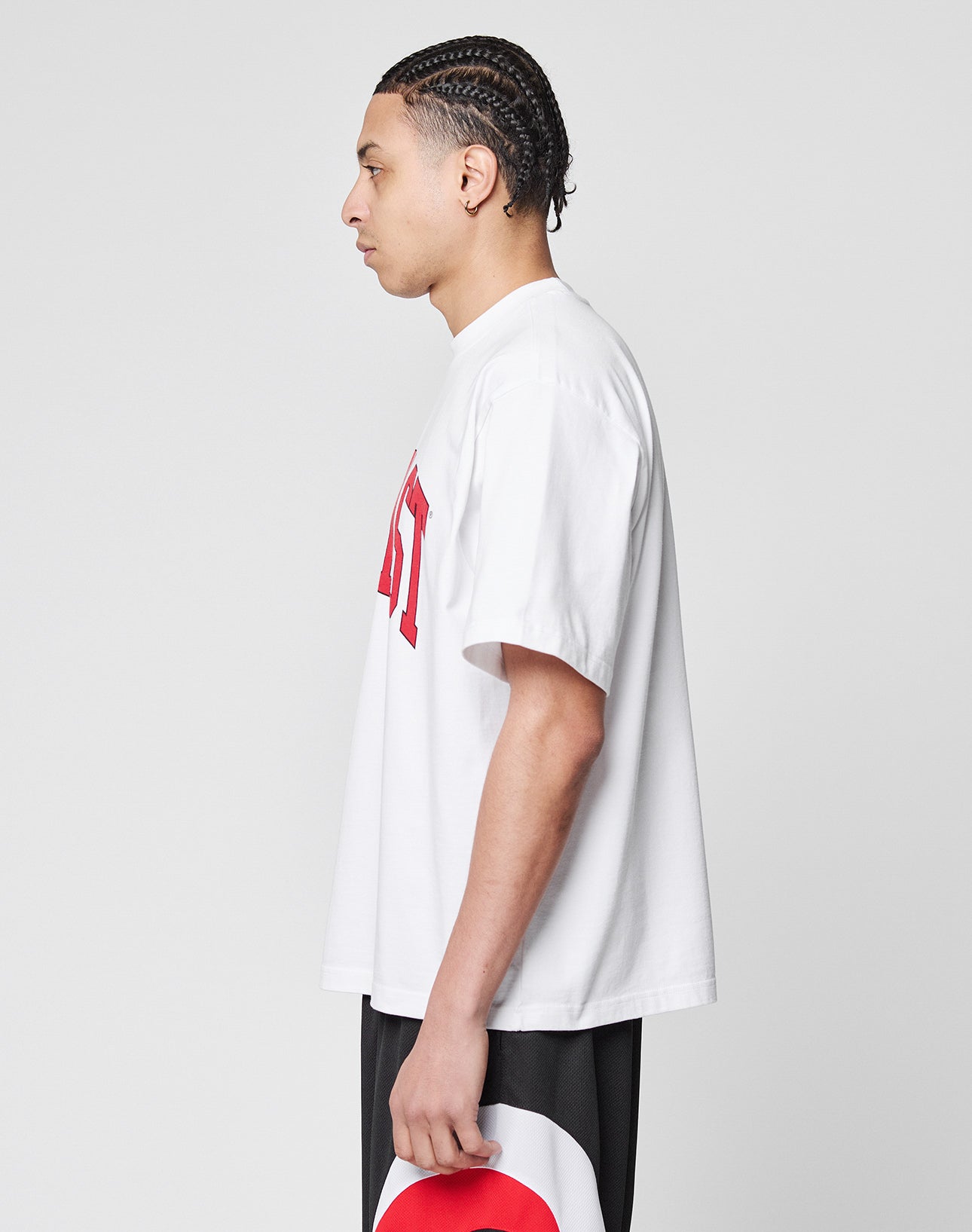 A person with braided hair stands in profile against a plain background, wearing the Live Fast College Tee—an oversize white T-shirt with red frontprint text—paired with black shorts and holding a white and red hat by their side.