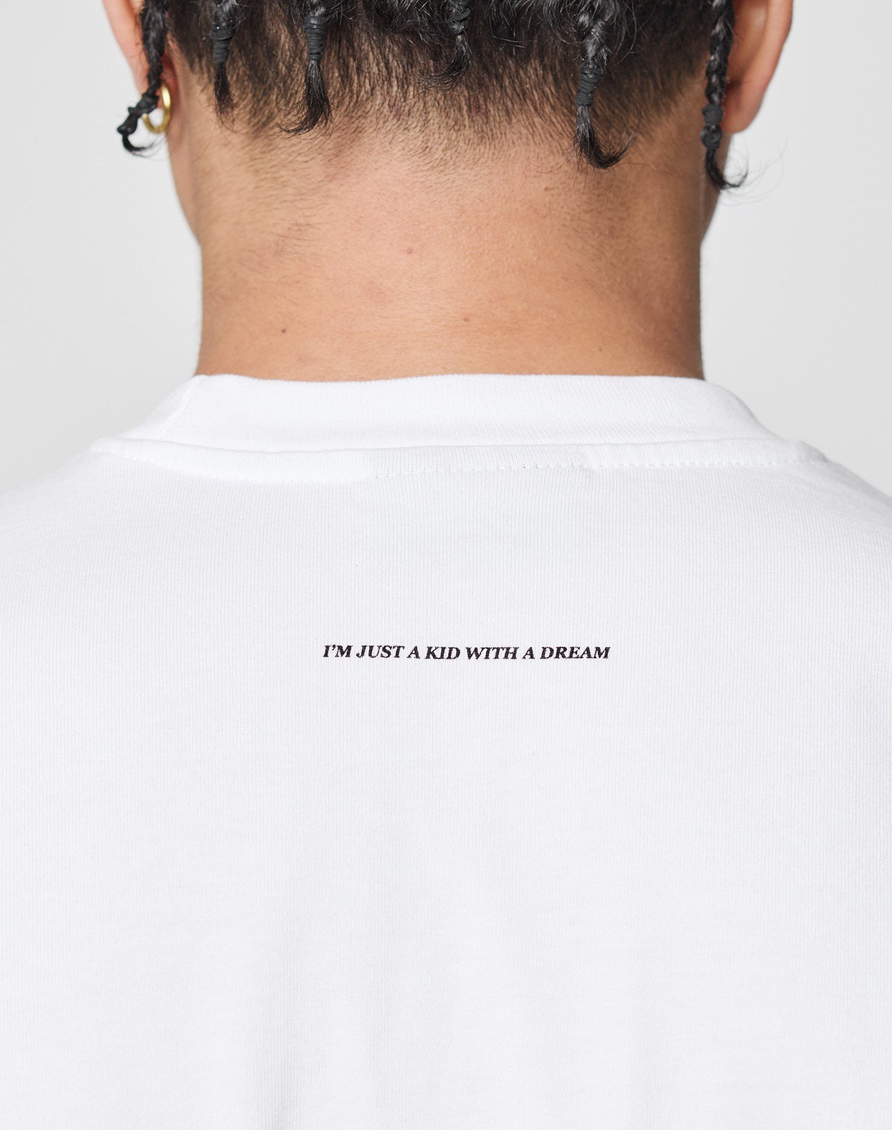 Close-up of the back of a persons neck and braided hair, wearing a Live Fast College Tee in Oversize Fit white 100% Baumwolle, with IM JUST A KID WITH A DREAM printed in small black letters on the upper back.