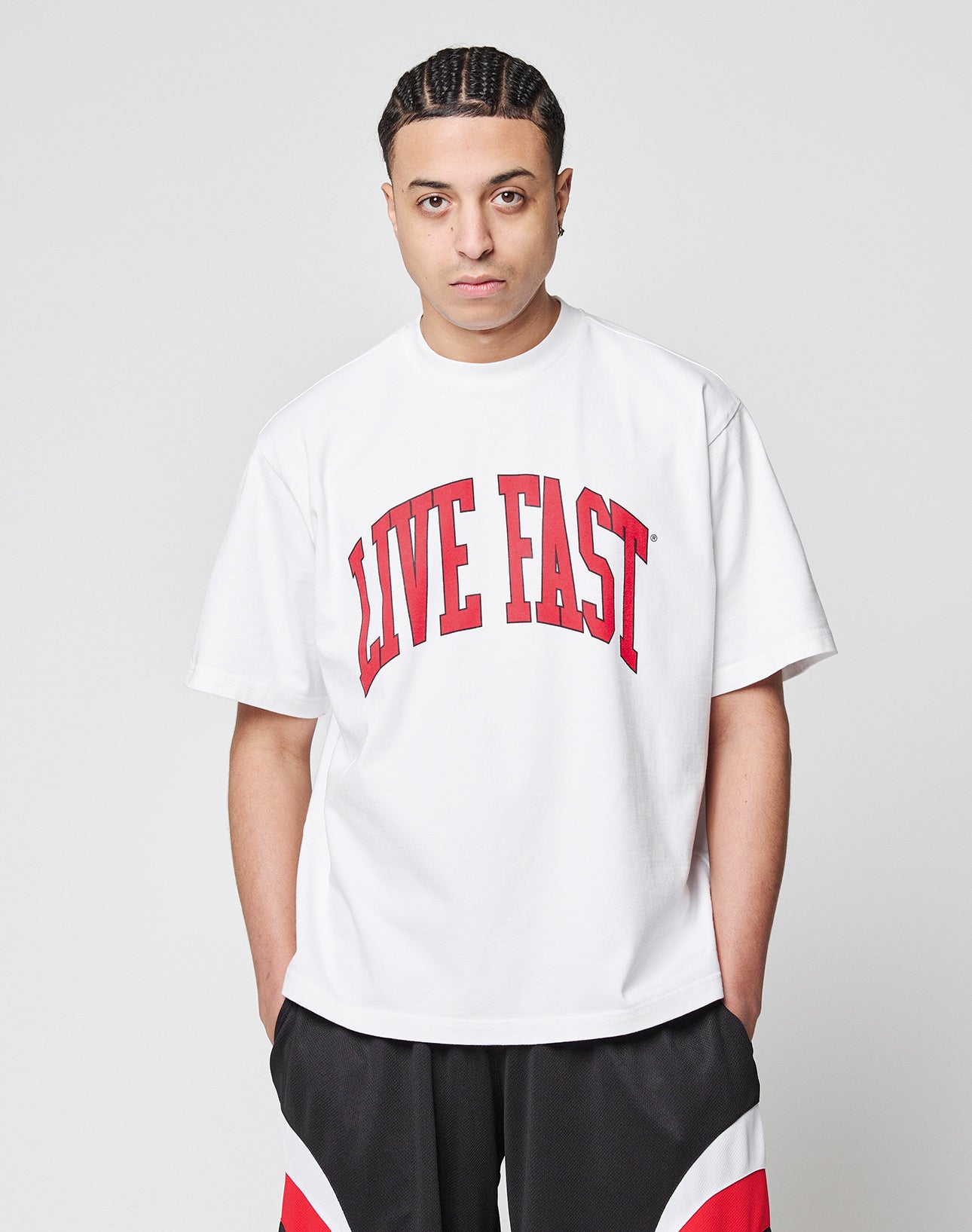 A person with short braided hair wears the LIVE FAST DIE YOUNG Live Fast College Tee, an oversize white t-shirt with bold red front letters, paired with black, white, and red shorts against a plain light gray background.