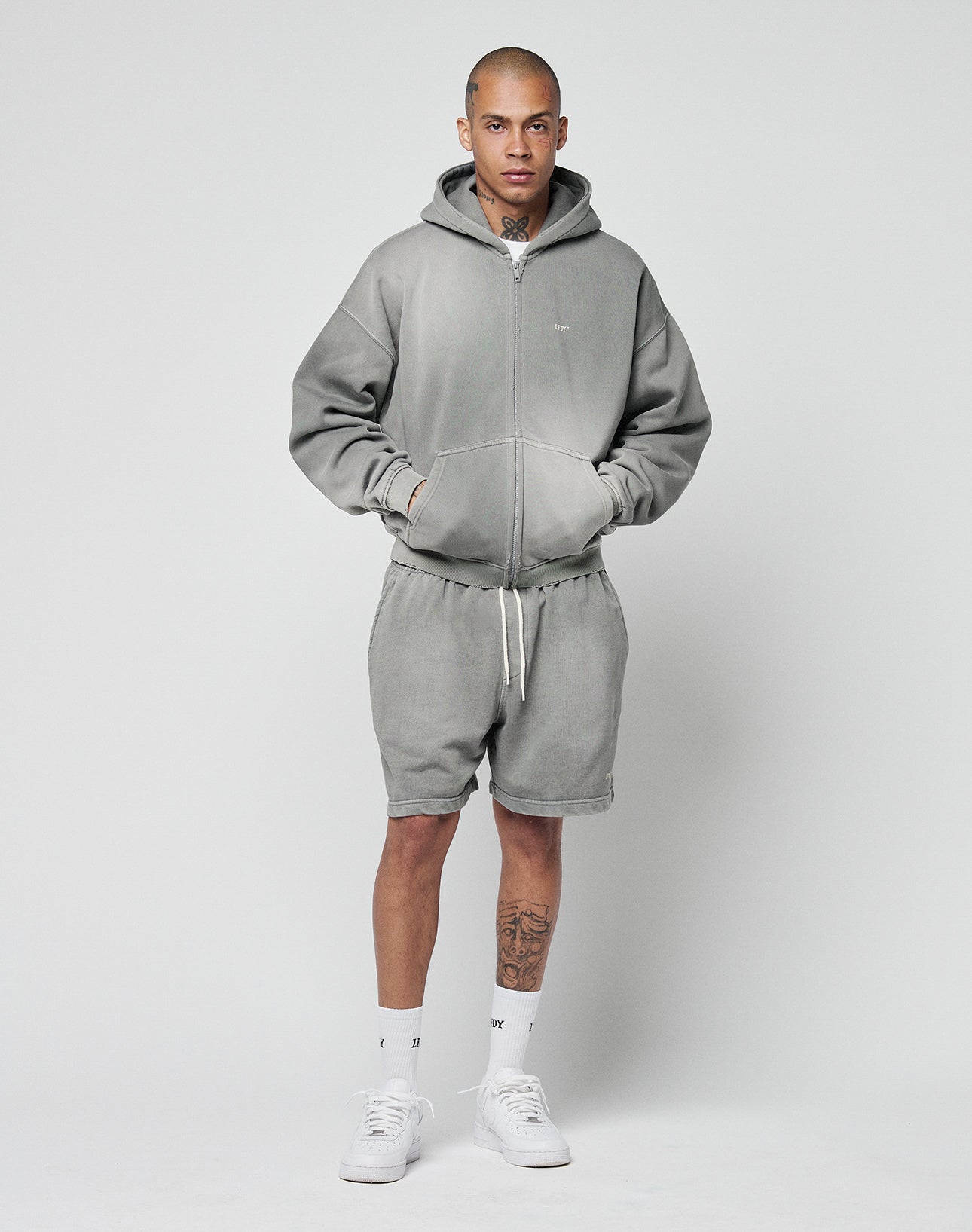 A person stands against a plain background wearing LIVE FAST DIE YOUNG Basic Sweat Shorts in grey, a matching regular fit zip-up hoodie with logo print, white socks, and white sneakers. Their hands are in their pockets and they look directly at the camera.