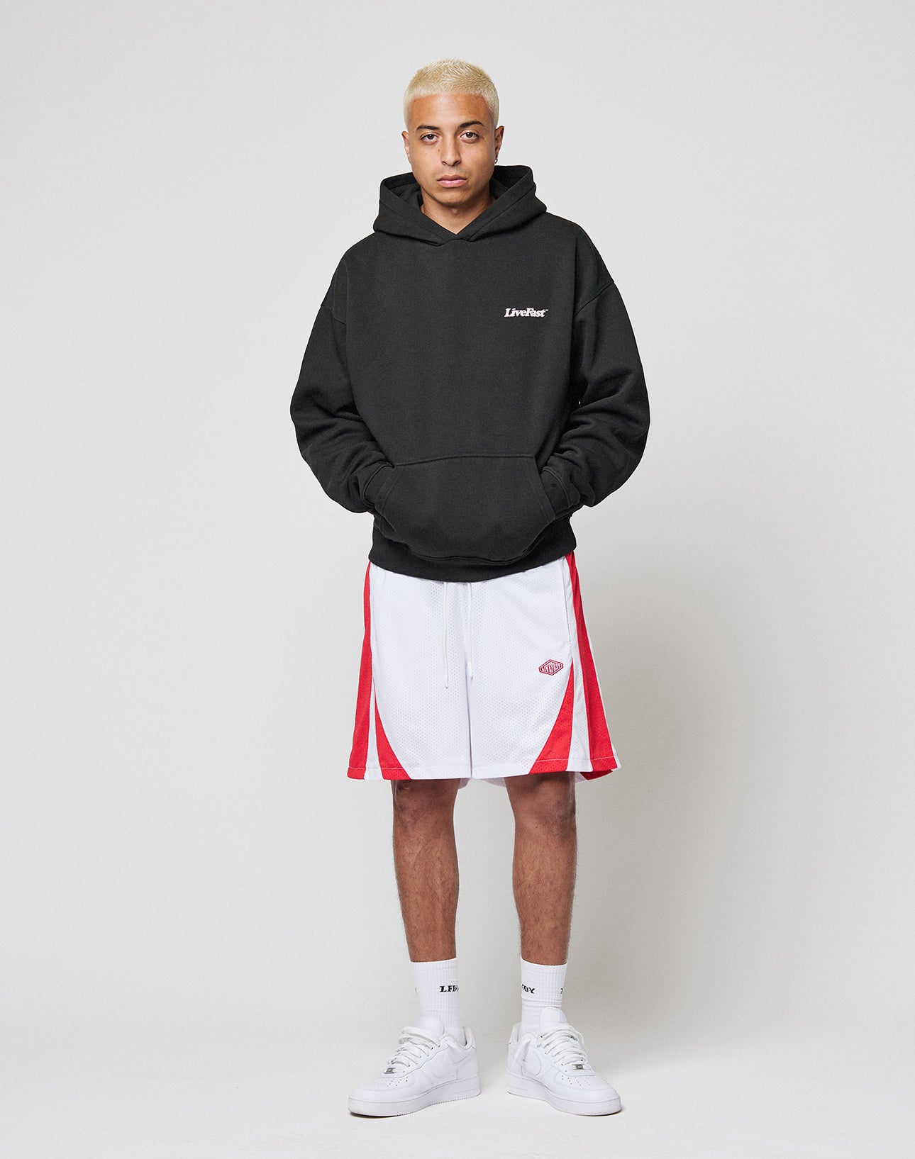 A person with short bleached blond hair wears a black LIVE FAST DIE YOUNG Basic 360 Hooded oversize hoodie, white and red athletic shorts, white socks, and white sneakers, standing against a plain light background.