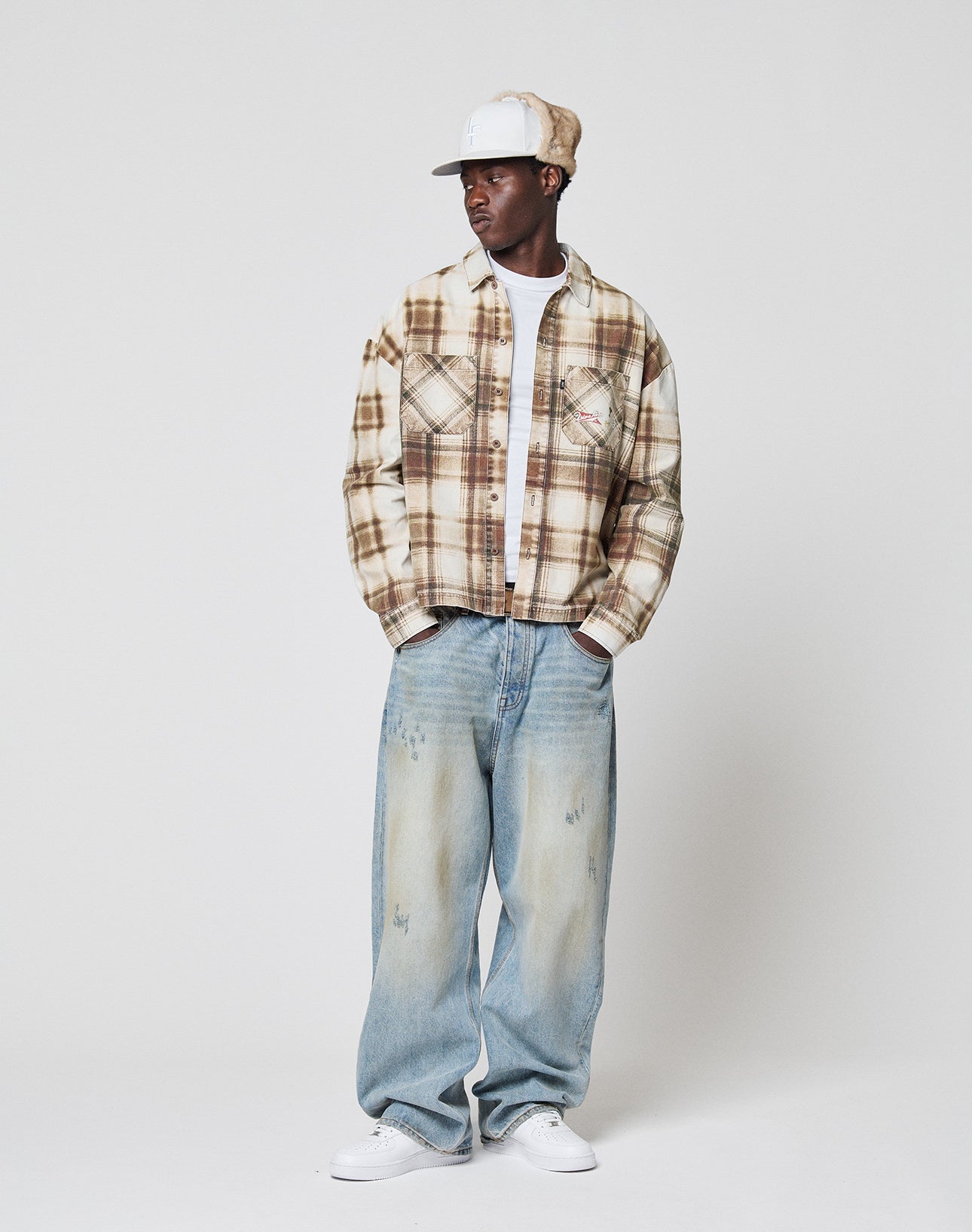 A man wearing a LIVE FAST DIE YOUNG Faded Flannel over a white t-shirt, paired with loose faded blue jeans, white sneakers, and a white cap stands against a plain white background.