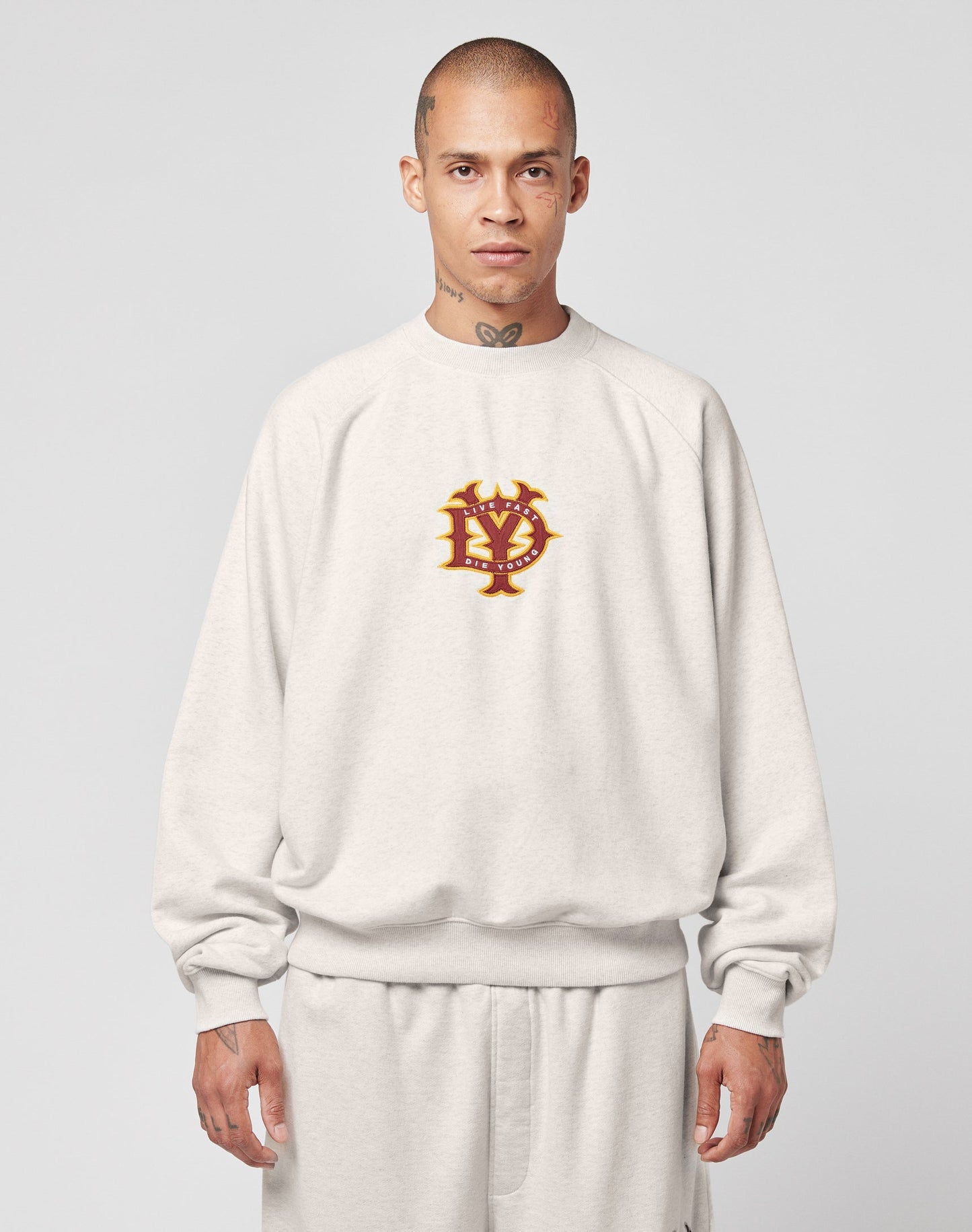 A person with a shaved head wears the LIVE FAST DIE YOUNG DY Sweater—an oversize, light gray sweatshirt with raglan sleeves and a red and gold interlocking letter emblem on the chest, paired with matching pants. The background is plain light gray.