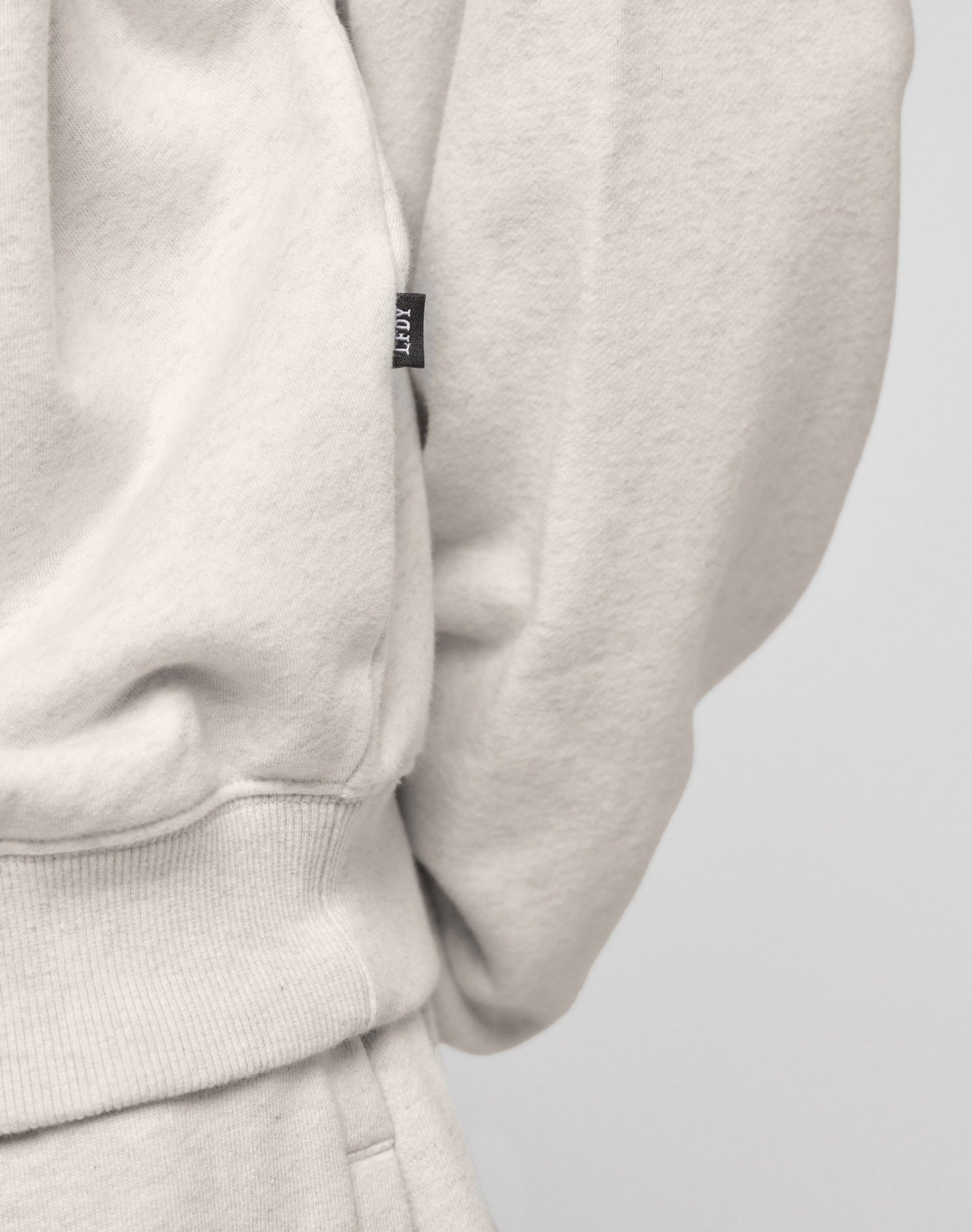 Close-up of a person wearing the DY Sweater by LIVE FAST DIE YOUNG in light beige with raglan sleeves, topstitching, a ribbed waistband, and a small black tag. The plain background highlights its modern Oversize Fit design.