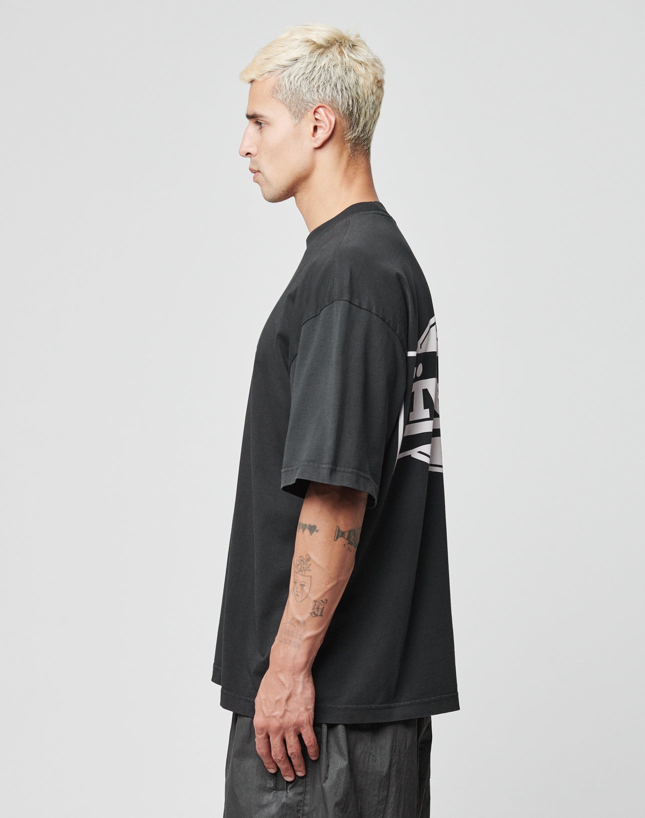 A person with short, bleached blond hair stands in profile, wearing the LIVE FAST DIE YOUNG LFDY Bulge Tee—an oversize black T-shirt with white front and back prints—and dark pants. Tattoos cover their left forearm. The background is plain light gray.