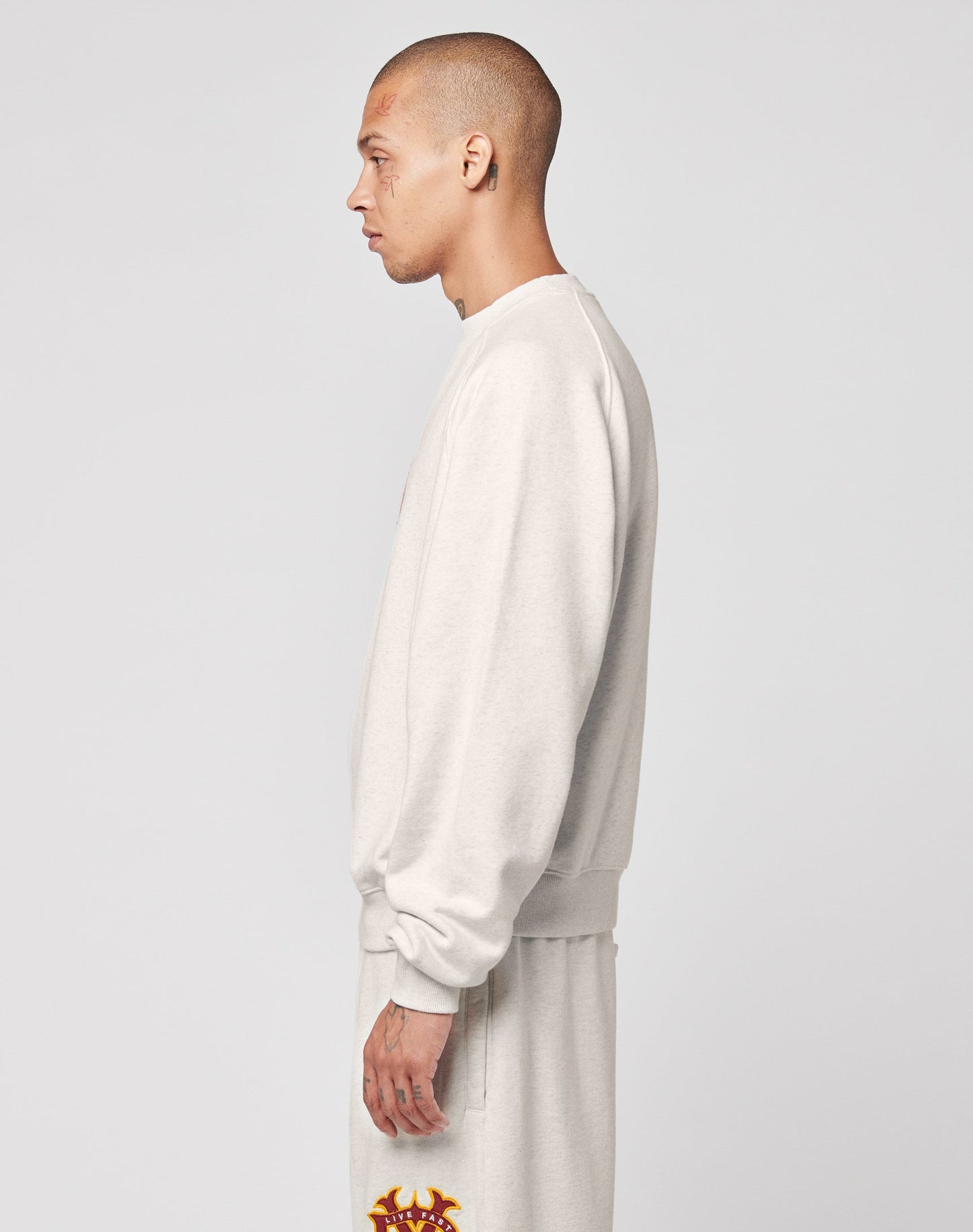 A man with closely shaved hair stands in profile, facing left, wearing the light gray DY Sweater and matching sweatpants by LIVE FAST DIE YOUNG with raglan sleeves and subtle topstitching, set against a plain light background.