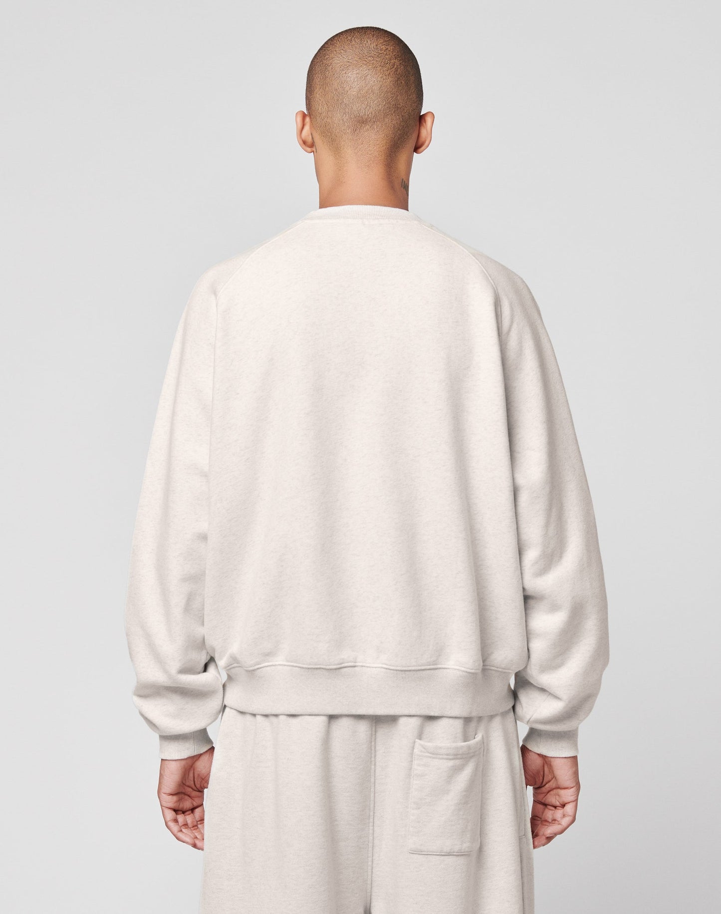 A person with a shaved head is seen from behind, wearing a light gray LIVE FAST DIE YOUNG DY Sweater with Raglan sleeves and matching pants against a plain, light-colored background.