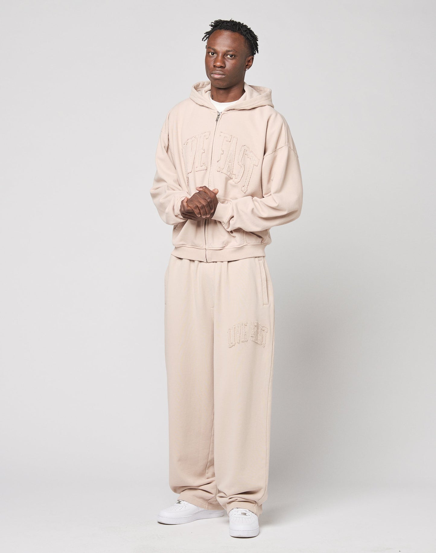 A person stands against a plain background wearing LIVE FAST DIE YOUNG’s Ripped College Wide Jogger in light beige cotton, paired with a matching wide hoodie and white sneakers. Embossed text adds detail as their hands are clasped in front.