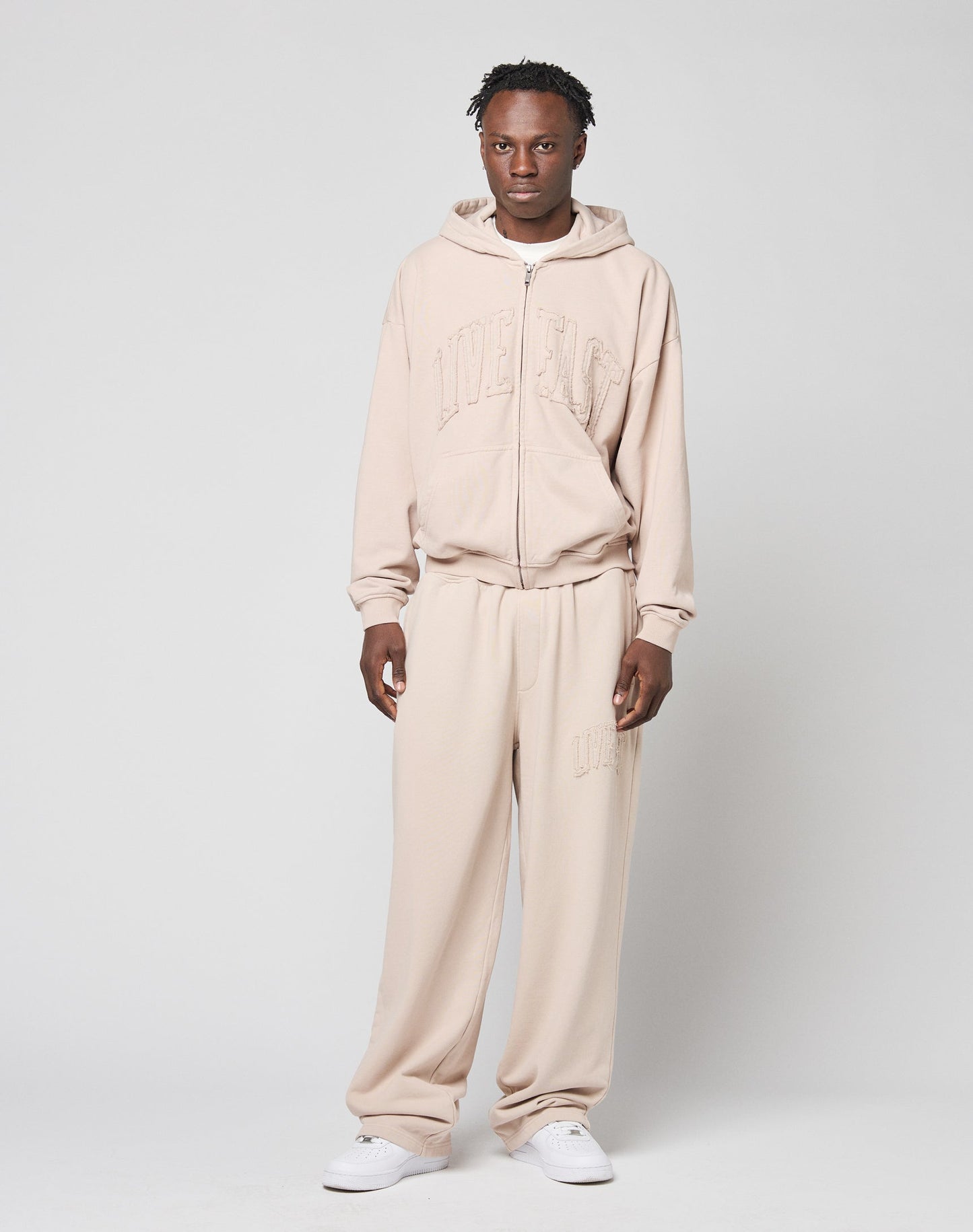 A person stands against a plain background wearing the oversize Ripped College Hooded Zip in light beige by LIVE FAST DIE YOUNG, made from weicher Baumwolle, paired with matching sweatpants and white sneakers. Subtle chest text is visible as they look at the camera.