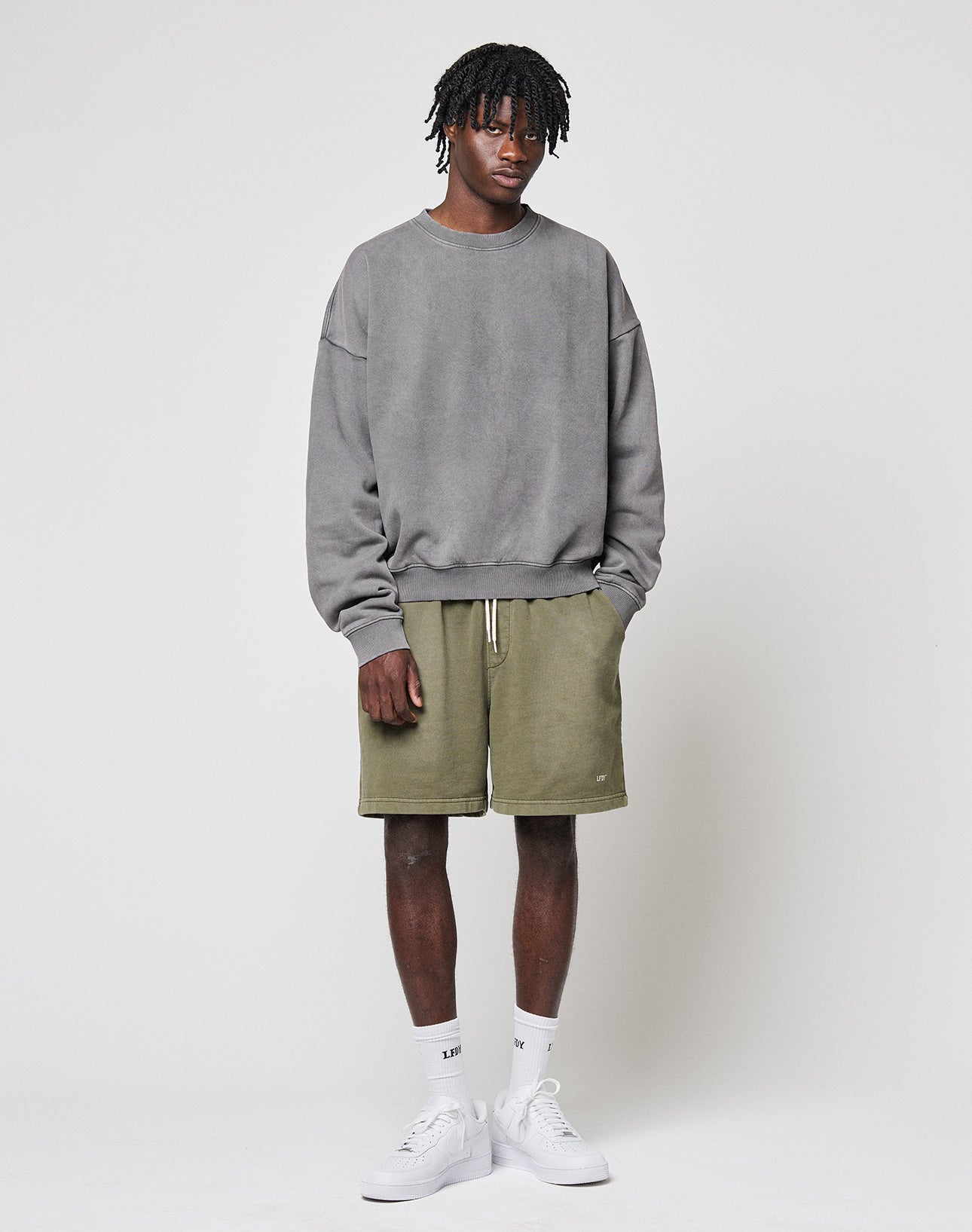 A man with short dreadlocks and a neutral expression wears the LIVE FAST DIE YOUNG Basic Blank Sweater in gray, paired with olive green shorts, white crew socks, and white sneakers against a plain white background.