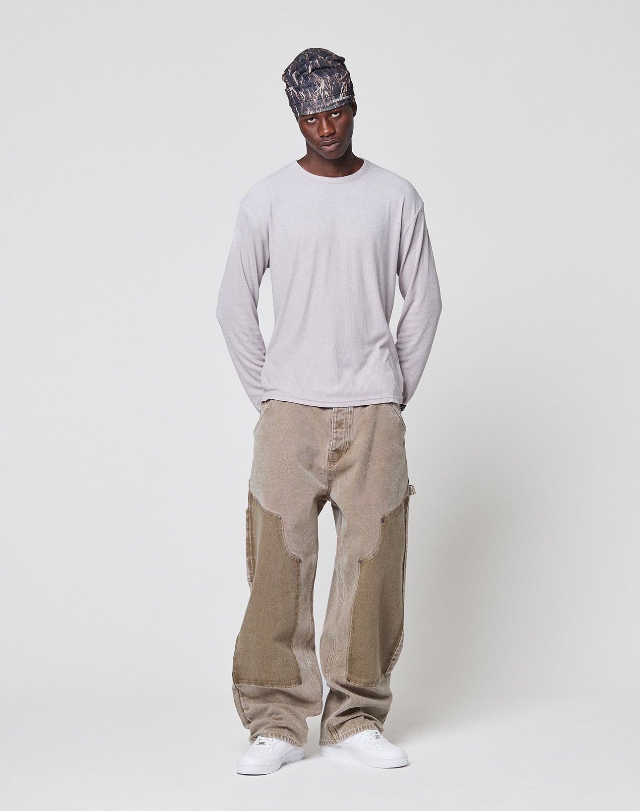 A man in a patterned headscarf, the LFDY Rib Longsleeve by LIVE FAST DIE YOUNG, oversized beige paneled jeans, and white sneakers stands against a plain white background with his hands behind his back.