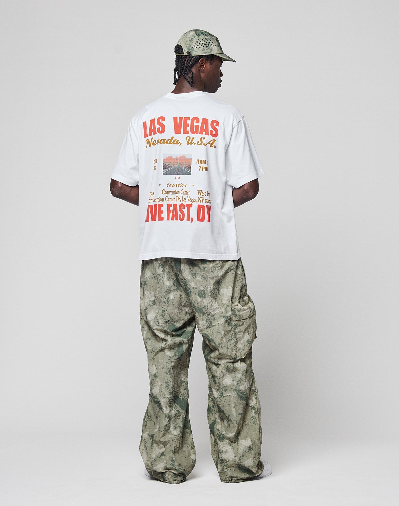 A person faces away, wearing the LIVE FAST DIE YOUNG Live Fast Vegas Tee (100% Baumwolle) in white with Las Vegas, Nevada, U.S.A. text, paired with green camouflage baggy pants and a matching cap against a plain light background.