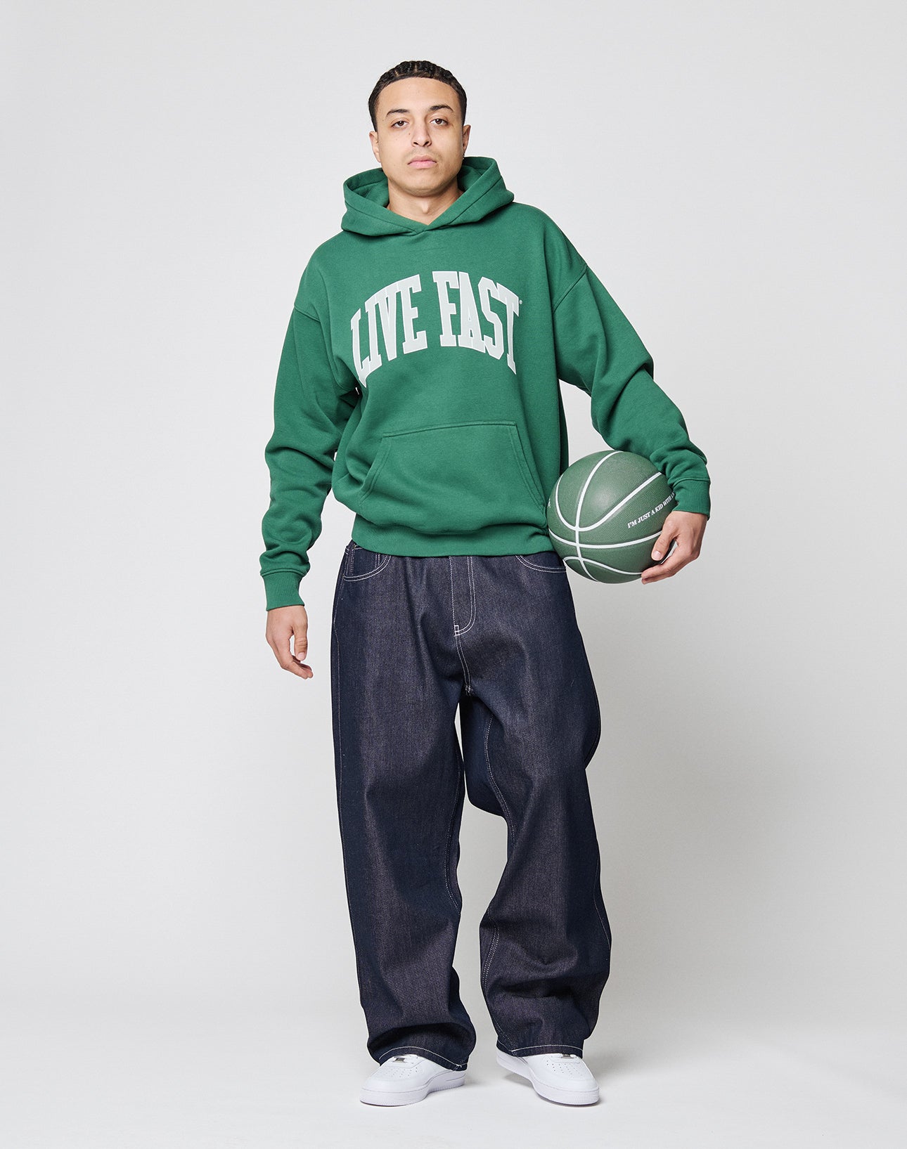 A person in a green Live Fast College Hooded by LIVE FAST DIE YOUNG with bold frontprint, baggy dark jeans, and white sneakers holds a green basketball against a plain light background.