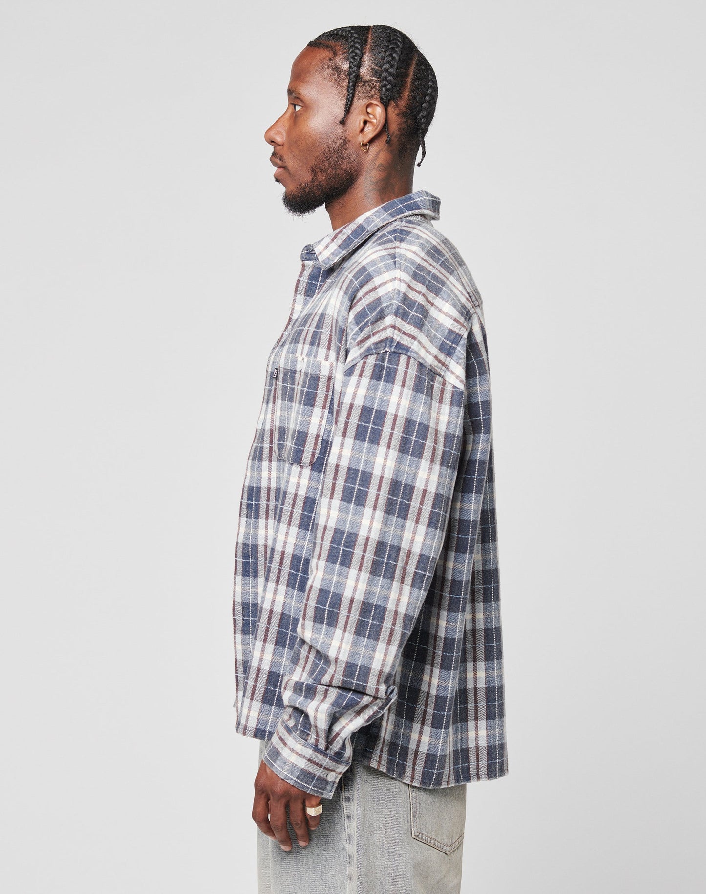 A man in profile against a plain background wears the LIVE FAST DIE YOUNG Vintage Flannel—a blue, white, and gray plaid oversize shirt with Brusttasche—paired with light jeans in Vintage Waschung.