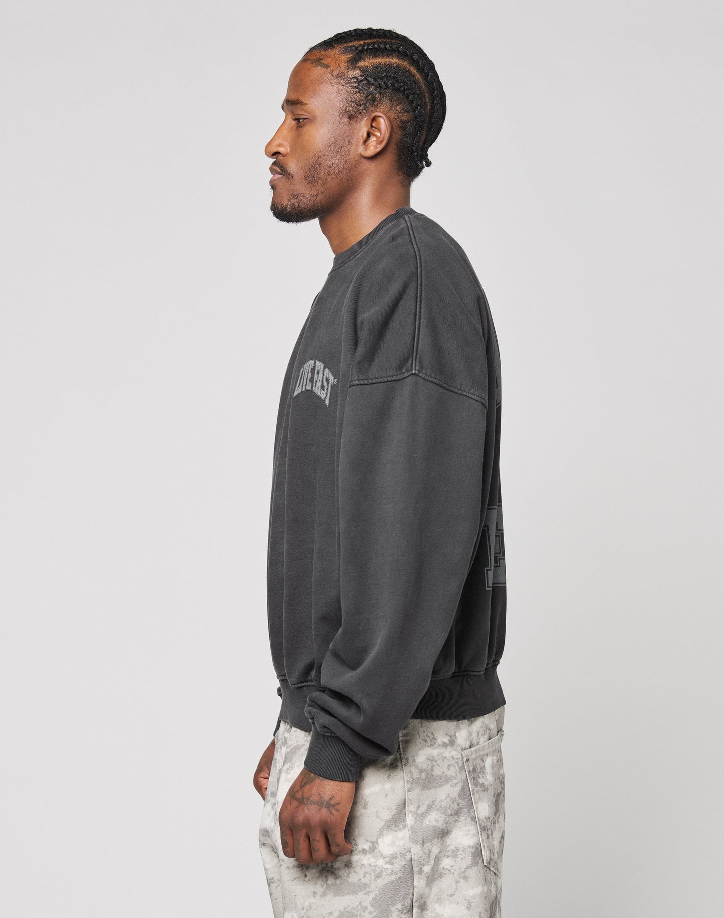 A man with braided hair stands in profile, wearing the LIVE FAST DIE YOUNG Live Fast Berlin Sweater—an oversized sweatshirt with topstitching—and light camo-patterned pants, set against a plain light background.