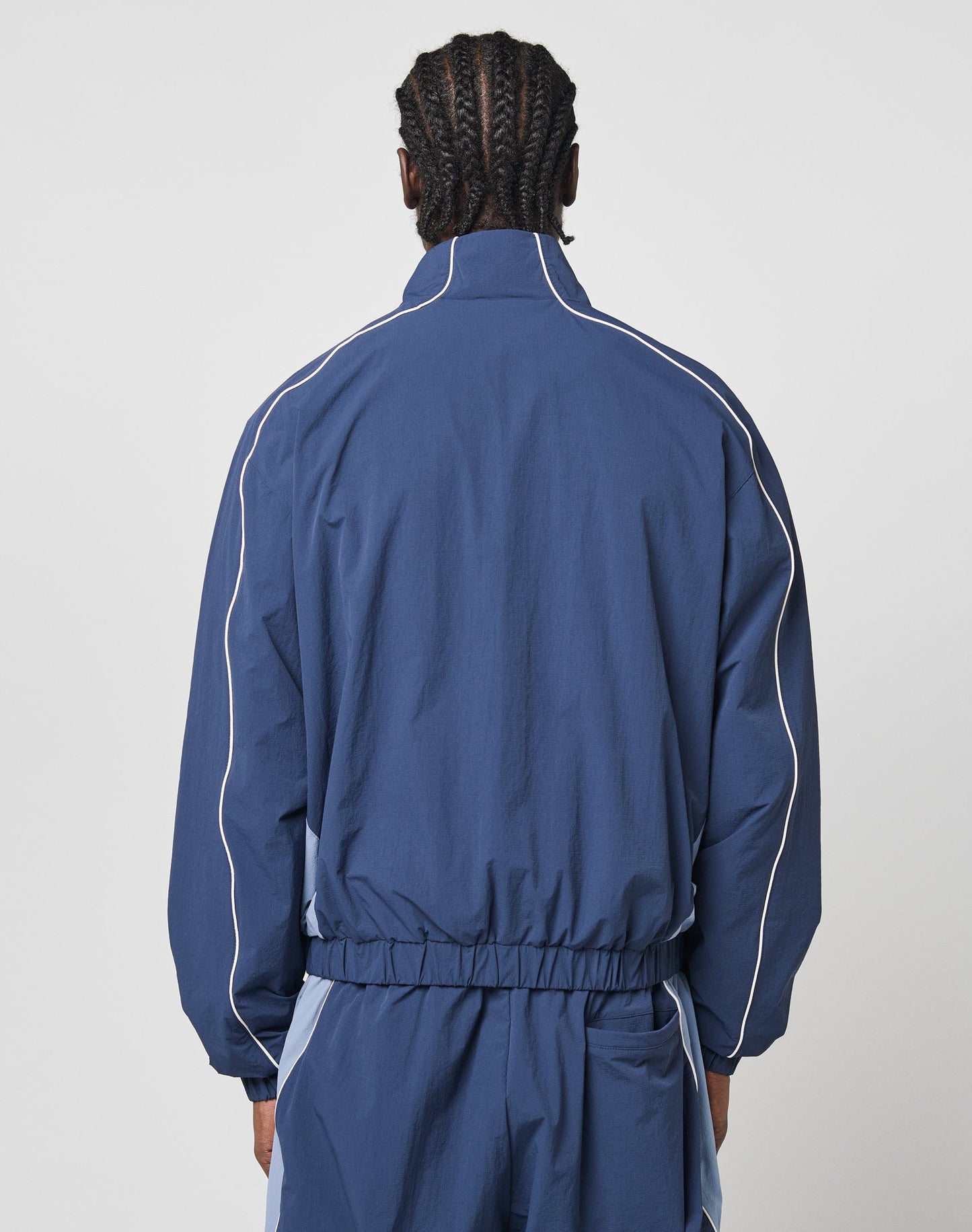 A person with braided hair wears the LIVE FAST DIE YOUNG Basic Nylon Trackjacket in navy blue, featuring white piping and elastic cuffs, viewed from behind against a plain light background.