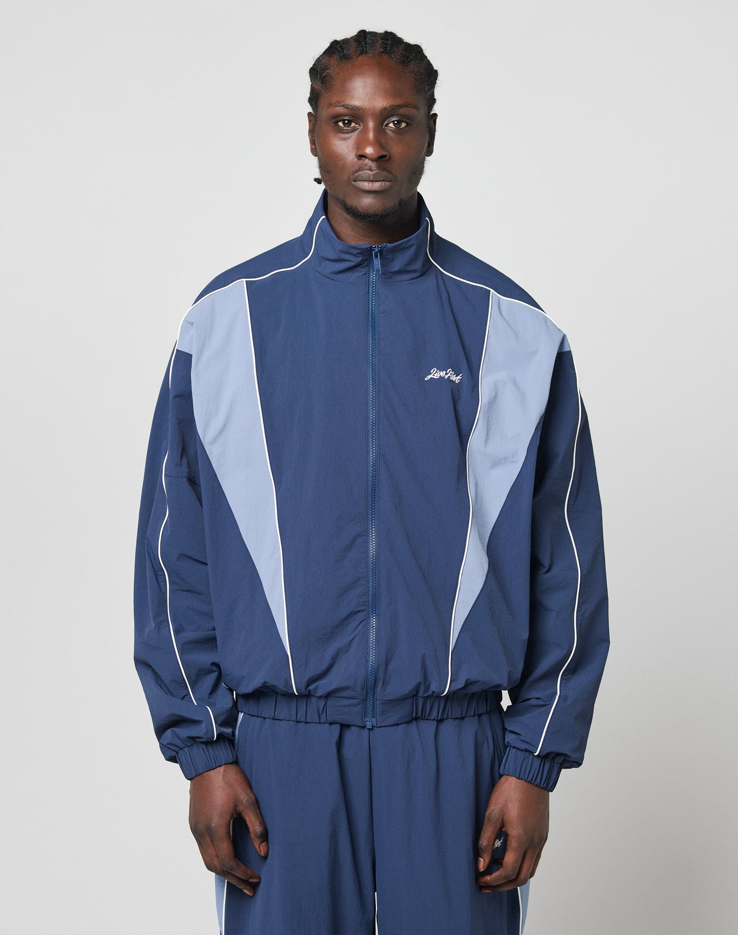 A person stands facing forward in a blue and light blue LIVE FAST DIE YOUNG Basic Nylon Trackjacket with matching pants and elastic cuffs, set against a plain light gray background.