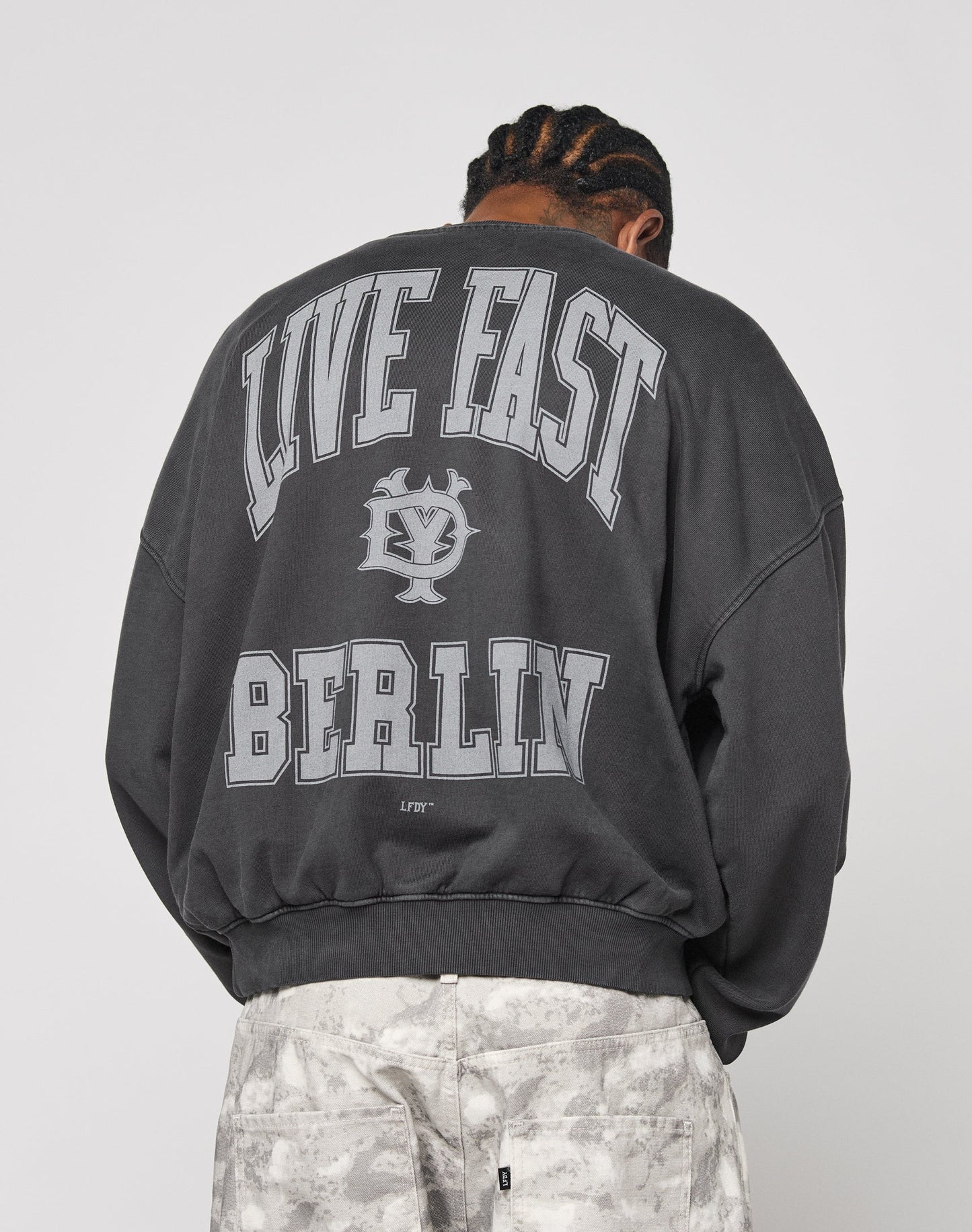 A person with braided hair faces away, wearing the LIVE FAST DIE YOUNG Live Fast Berlin Sweater—an oversized dark sweatshirt with bold white “LIVE FAST BERLIN” text and a central graphic—paired with patterned light pants and topstitching details.