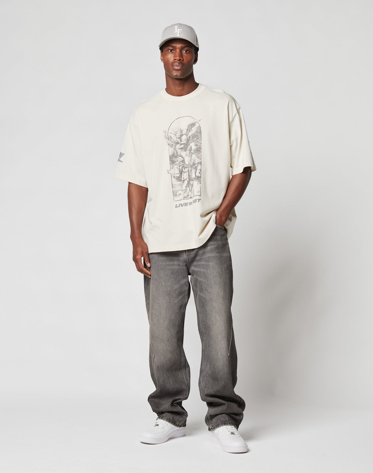 A man stands facing forward with one hand in his pocket, wearing a light gray baggy baseball cap, an oversized off-white 100% Baumwolle graphic T-shirt, LIVE FAST DIE YOUNG Rhomb Wide Denim faded gray jeans, and white sneakers.