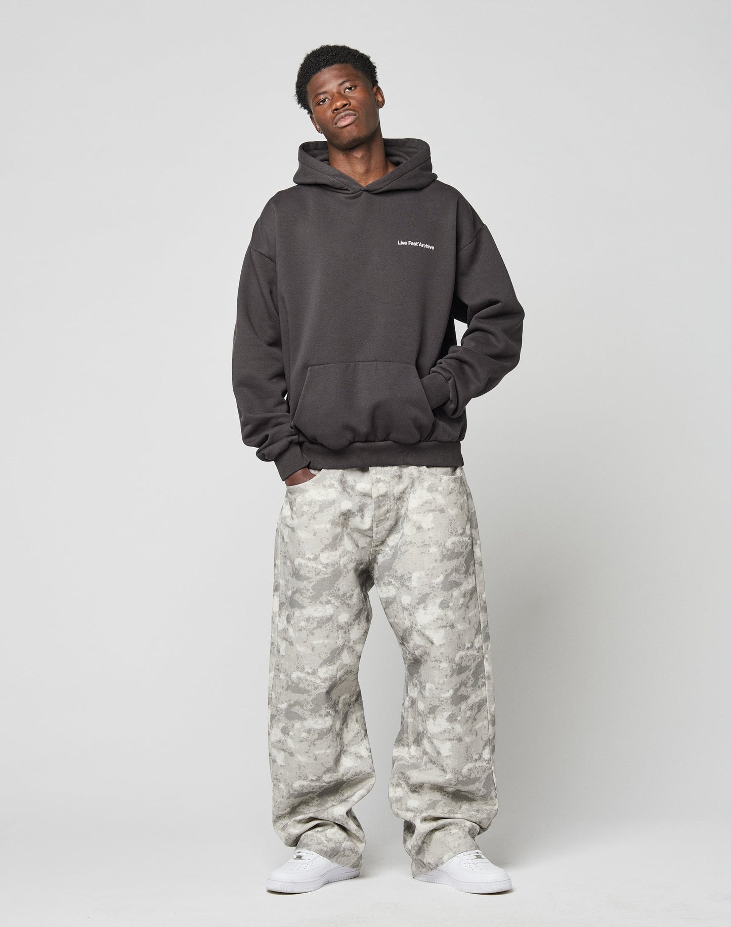 A young man stands against a plain background wearing the LIVE FAST DIE YOUNG LFDY Middlefinger Hooded, light gray camouflage cargo pants, and white sneakers, hands in his pockets and a relaxed expression.