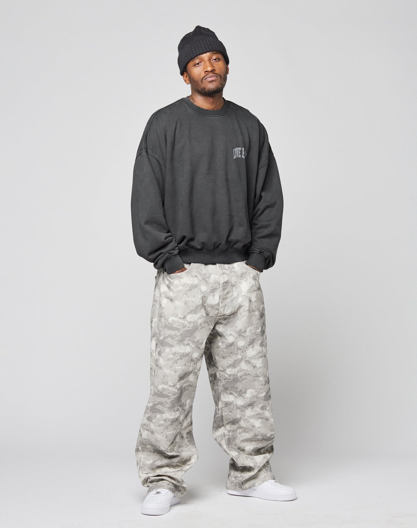 A man stands relaxed against a plain light gray background, wearing the LIVE FAST DIE YOUNG Live Fast Berlin Sweater in dark hues, paired with a black beanie, baggy gray camo pants, and white sneakers. Visible topstitching accents the oversized fit.