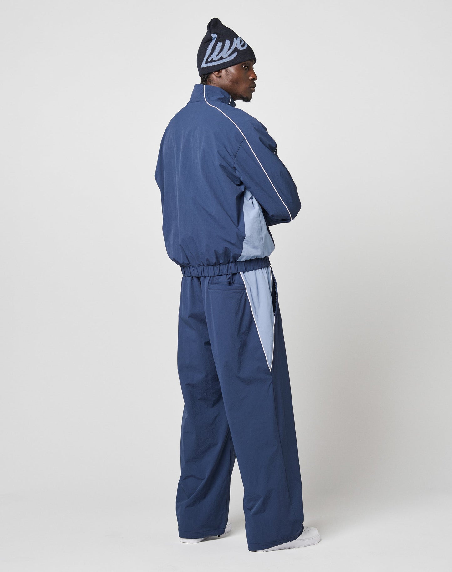 A person in a navy blue LIVE FAST DIE YOUNG Basic Nylon Trackjacket with light blue accents, elastic cuffs, and a navy beanie stands in a relaxed sideways pose against a plain white background.