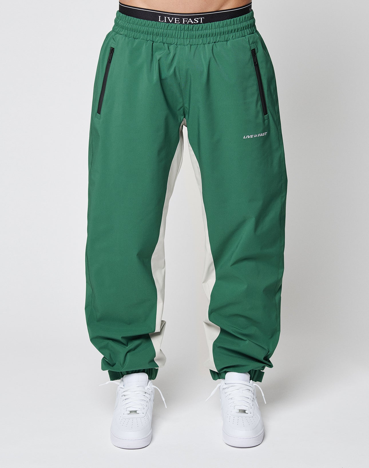 A person wears green and white LIVE FAST DIE YOUNG Basic Shell Pants with an elastic drawstring waist, black zip pockets, LIVE FAST logos on waistband and leg, paired with white sneakers against a plain light background.