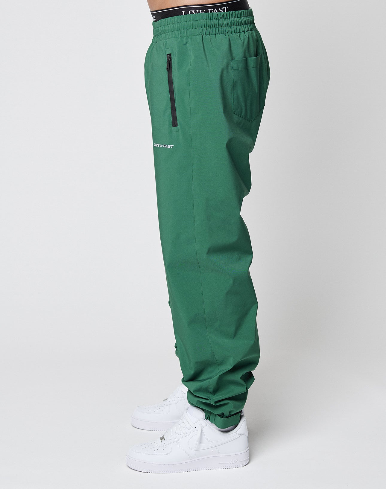 A person stands sideways against a plain white background wearing LIVE FAST DIE YOUNG Basic Shell Pants in green with an elastic waistband, drawcord, black zipper pocket, reflective LIVE FAST thigh print, and white sneakers.