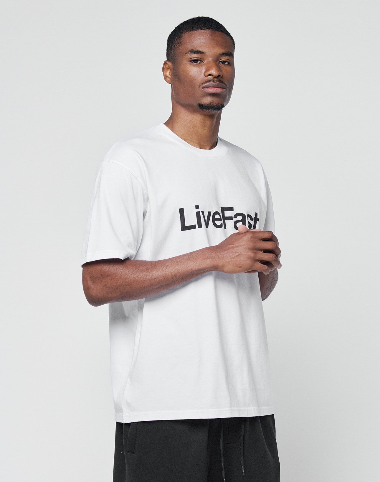 A man stands against a white background wearing the LIVE FAST DIE YOUNG Basic LiveFast Tee—a regular fit white t-shirt with “LiveFast” in bold black letters—and black pants, looking neutrally at the camera.