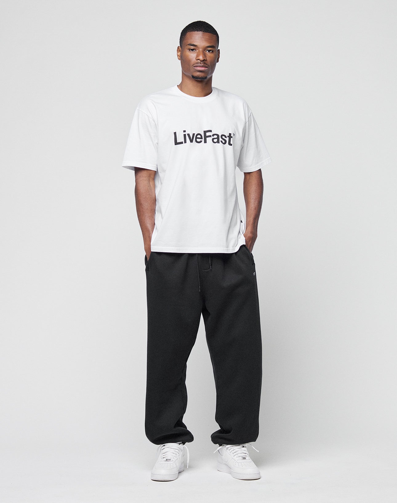 A man faces forward in a white LIVE FAST DIE YOUNG Basic LiveFast Tee with LiveFast in black, paired with loose black pants and white sneakers, against a plain light background.