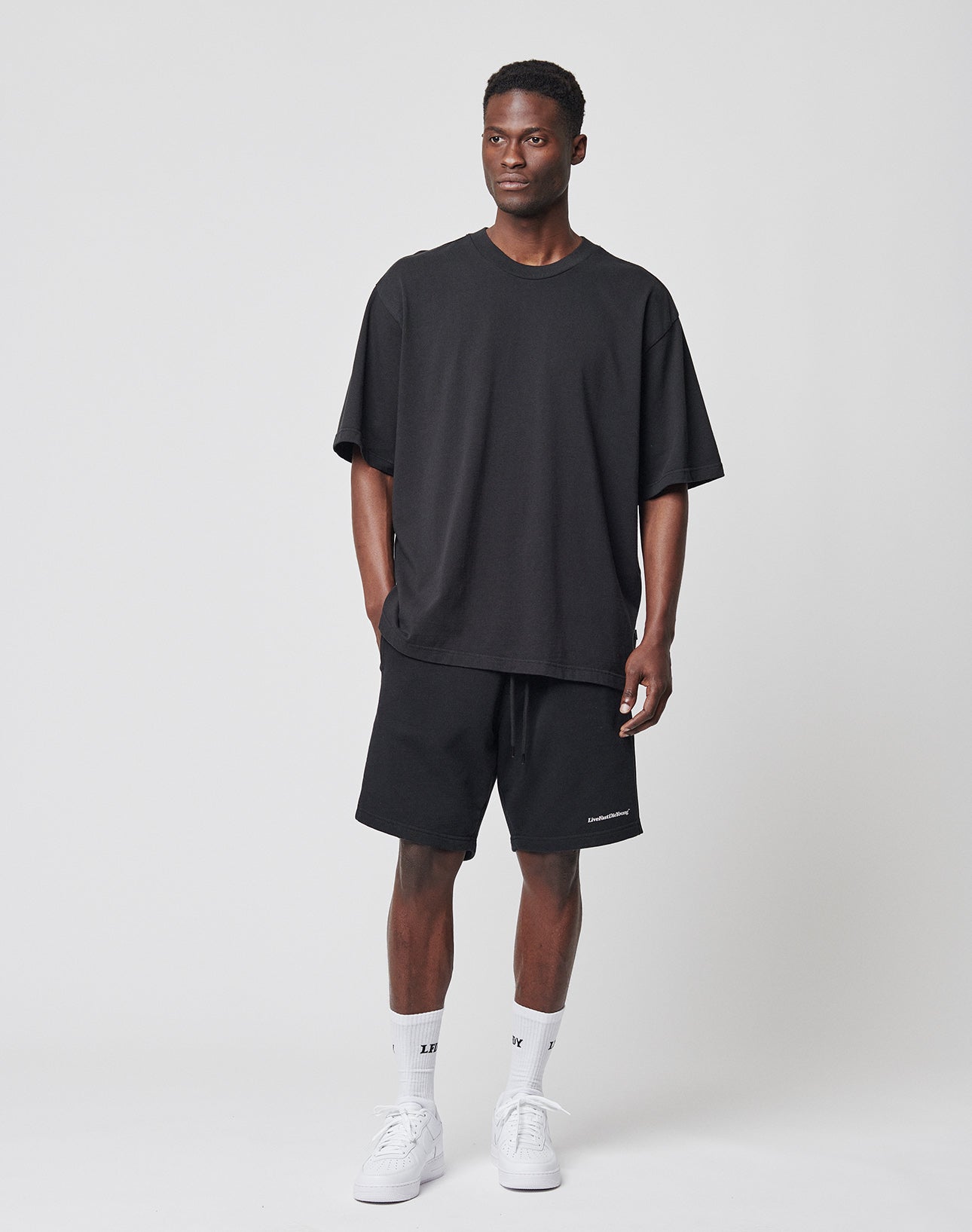 A man stands against a plain white background wearing a loose black LIVE FAST DIE YOUNG Basic Heavy Tee, black shorts, white crew socks, and white sneakers. He has short hair, relaxed hands at his sides, and care instructions on the tag.