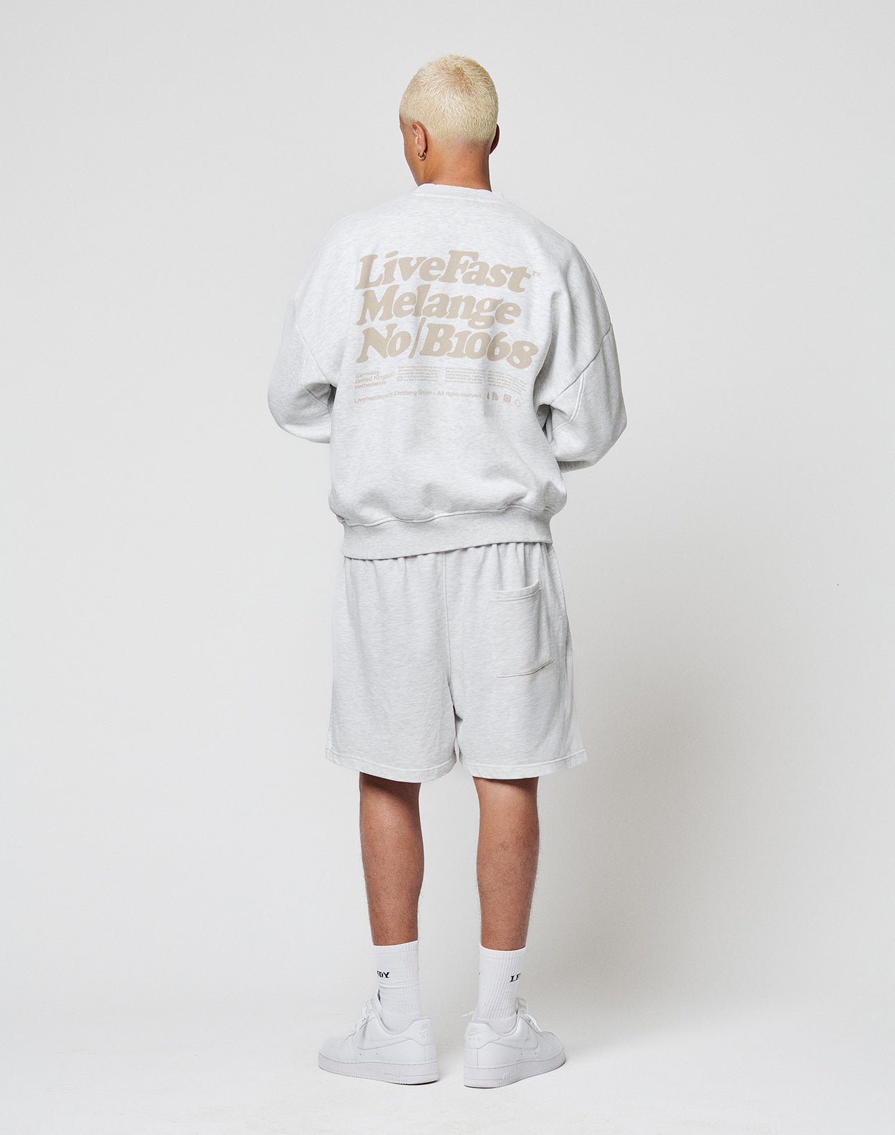 A person with short blond hair, wearing a light gray Basic Melange Sweater by LIVE FAST DIE YOUNG and matching shorts, stands facing away. The outfit is paired with white socks and sneakers.