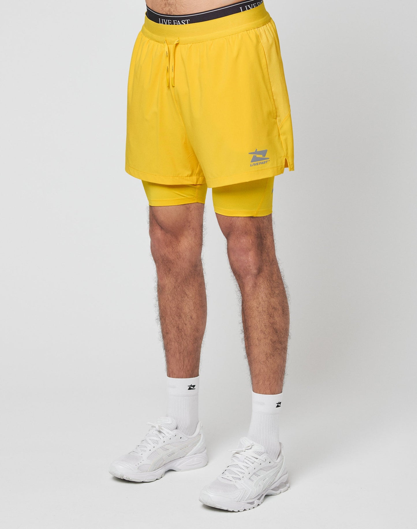 A person wearing LIVE FAST DIE YOUNG Live Fast Performance Running Shorts in yellow, featuring integrated tights, along with white socks and sneakers stands against a plain white background. Only the lower body is visible.