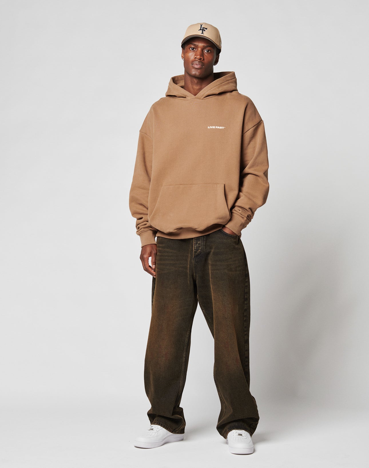 A man wears the LIVE FAST DIE YOUNG Live Fast Plain Hooded tan hoodie with dark baggy jeans, white sneakers, and a beige baseball cap, standing against a plain light gray background.