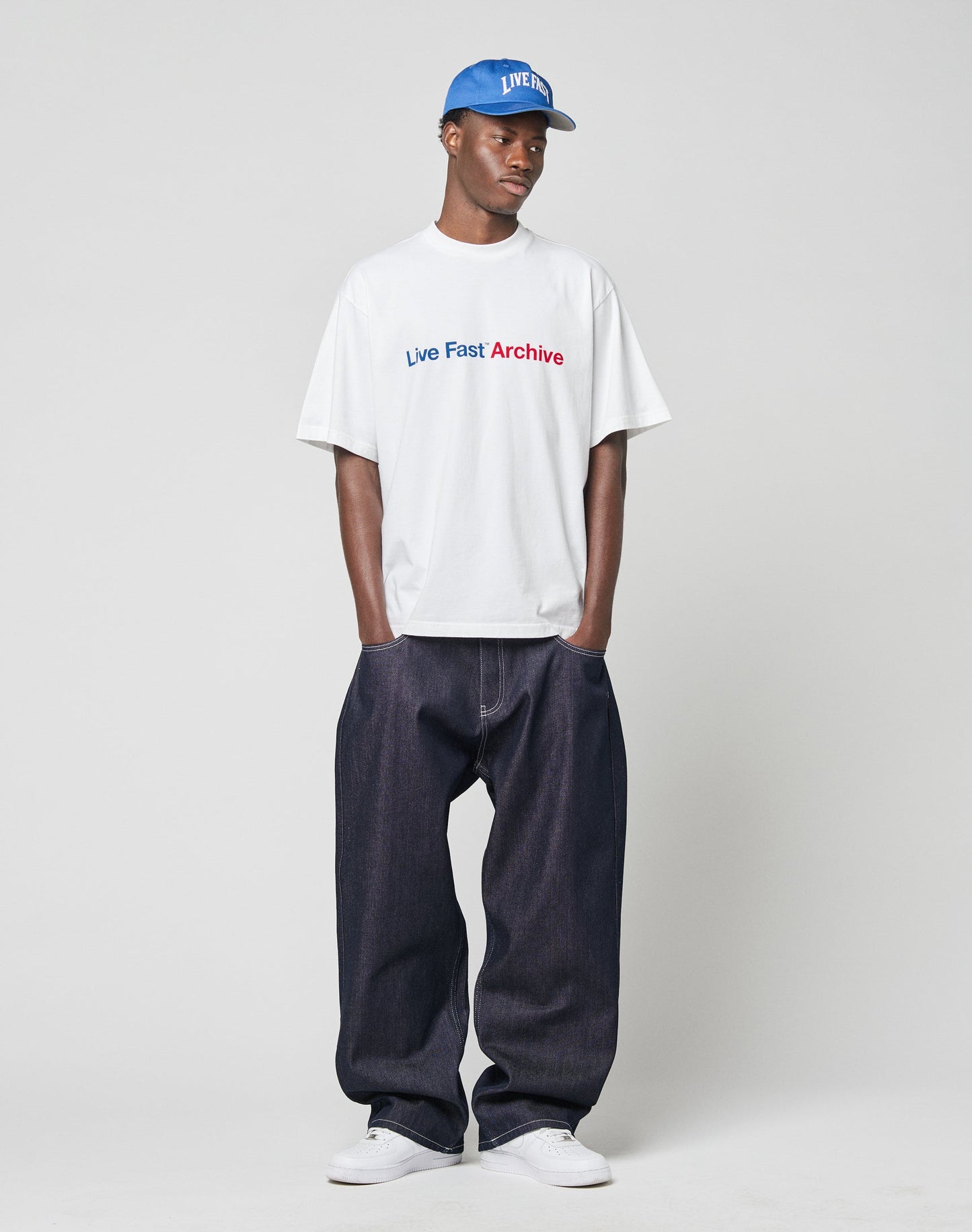 A man in a blue cap, white LIVE FAST DIE YOUNG Live Fast Archive Tee, baggy dark jeans, and white sneakers stands against a plain light background with his hands in his pockets.