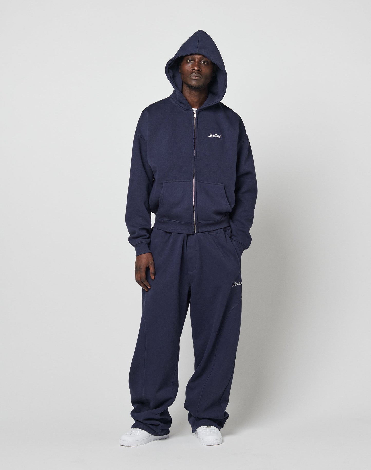 A person stands against a plain background wearing LIVE FAST DIE YOUNGs Basic Script Hooded Zip in navy blue with the hood up and matching sweatpants, both featuring small white logos and a branded zipper puller, paired with white sneakers.