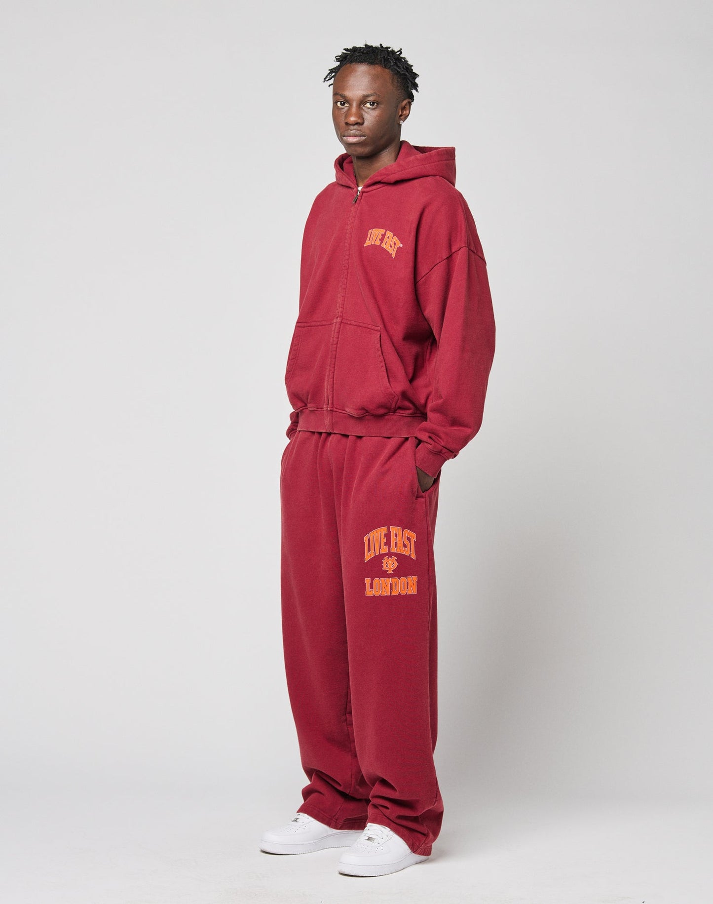 A person stands against a plain background wearing LIVE FAST DIE YOUNG’s Live Fast London Wide Jogger set in maroon with yellow text, Elastikbund waist, hands in pockets, and white sneakers.