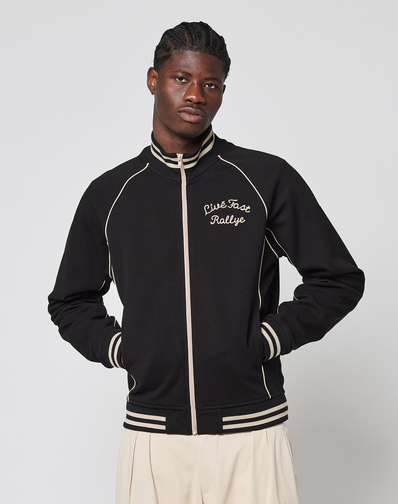 A man stands against a plain light gray background wearing the Live Fast Tracksuit Jacket—a black zip-up with beige trim, Chainstitch Logo, Cise Fast Ralye text on the left chest, and hands in the Eingrifftaschen.