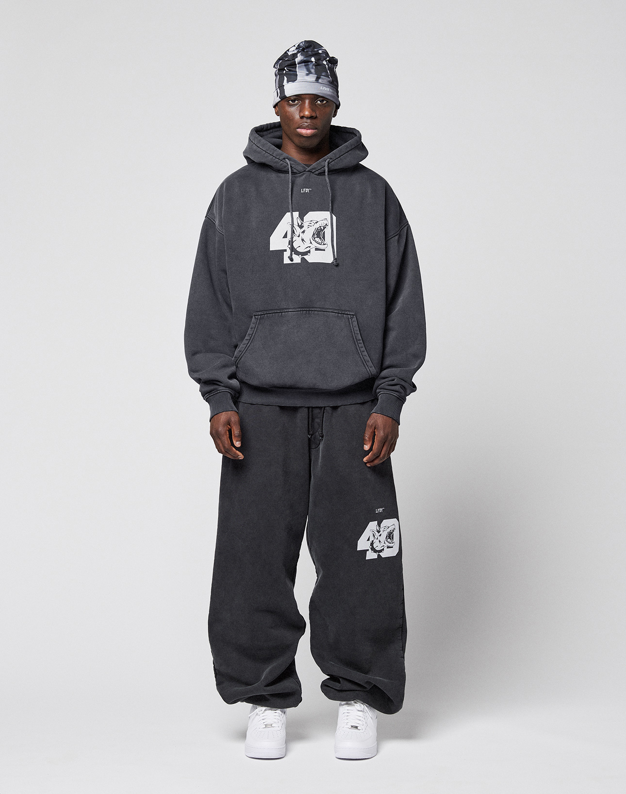 A man faces forward wearing the LIVE FAST DIE YOUNG LFDY 40 Hooded in dark gray with white graphics and a Kangaroo-Tasche, paired with matching baggy sweatpants, a patterned headscarf, and white sneakers against a plain light gray background.