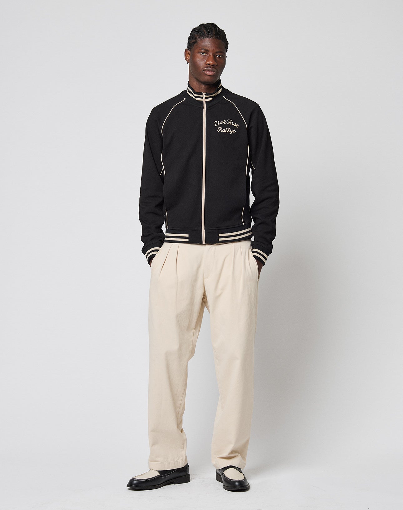 A man faces forward against a plain light background, wearing the Live Fast Tracksuit Jacket in black with white trim and Eingrifftaschen, paired with cream Regular Fit pleated pants and black-and-white shoes.