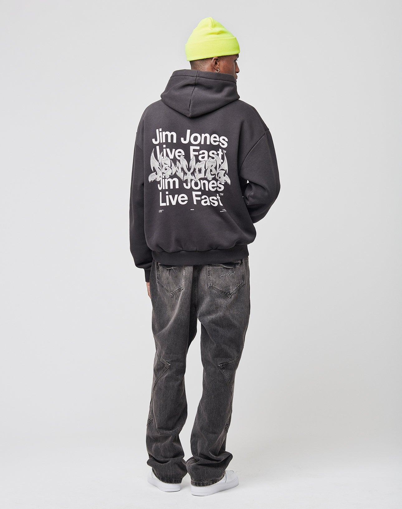 A person faces away, wearing a neon yellow beanie, loose dark-wash jeans, white sneakers, and the LIVE FAST DIE YOUNG Live Fast NY Hooded—an oversize black hoodie with Jim Jones front and back print, made from 70% Baumwolle.