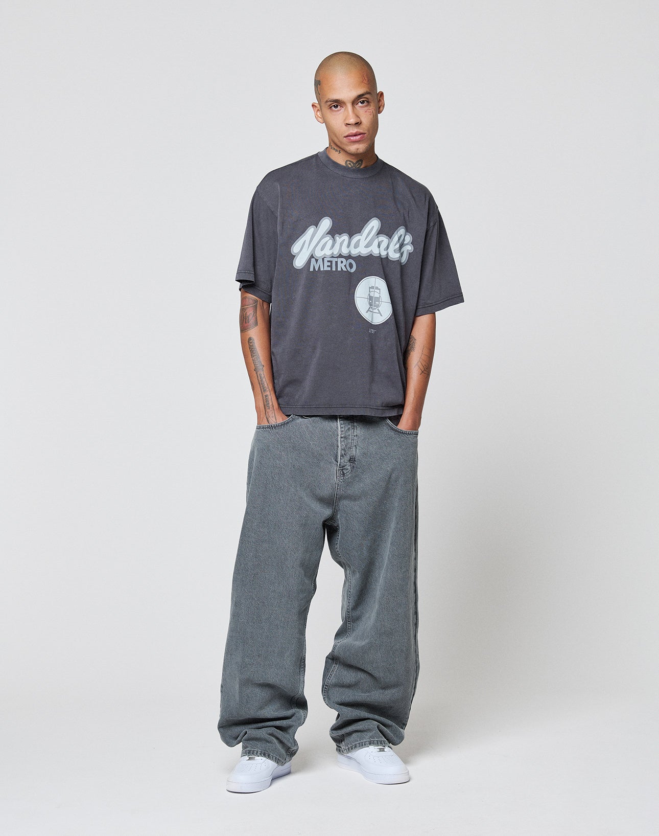 A man with a shaved head stands facing forward, hands in pockets, wearing the LIVE FAST DIE YOUNG LFDY Vandals Tee in dark gray and baggy gray jeans. The oversized 100% cotton shirt pairs perfectly with white sneakers against a plain light background.