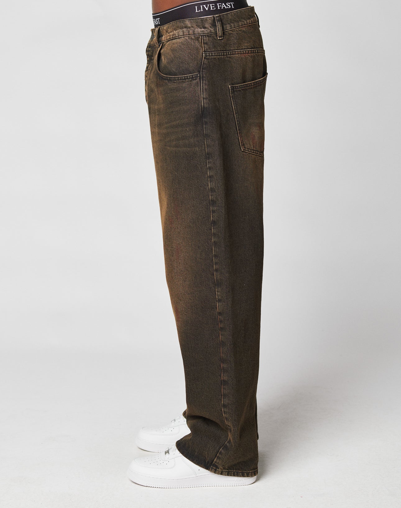 A person stands sideways in LFDY Baggy Denim by LIVE FAST DIE YOUNG—faded brown, 100% cotton jeans with a flag label. Black LIVE FAST waistband underwear peeks out above white sneakers, set against a plain, light-colored background.