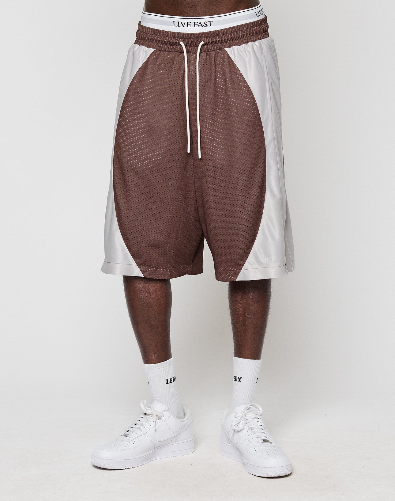 Wearing LIVE FAST DIE YOUNG LFDY League Shorts in brown and white mesh with a logo-print waistband, paired with white LIVE FAST crew socks and white sneakers, a person stands against a plain light background.
