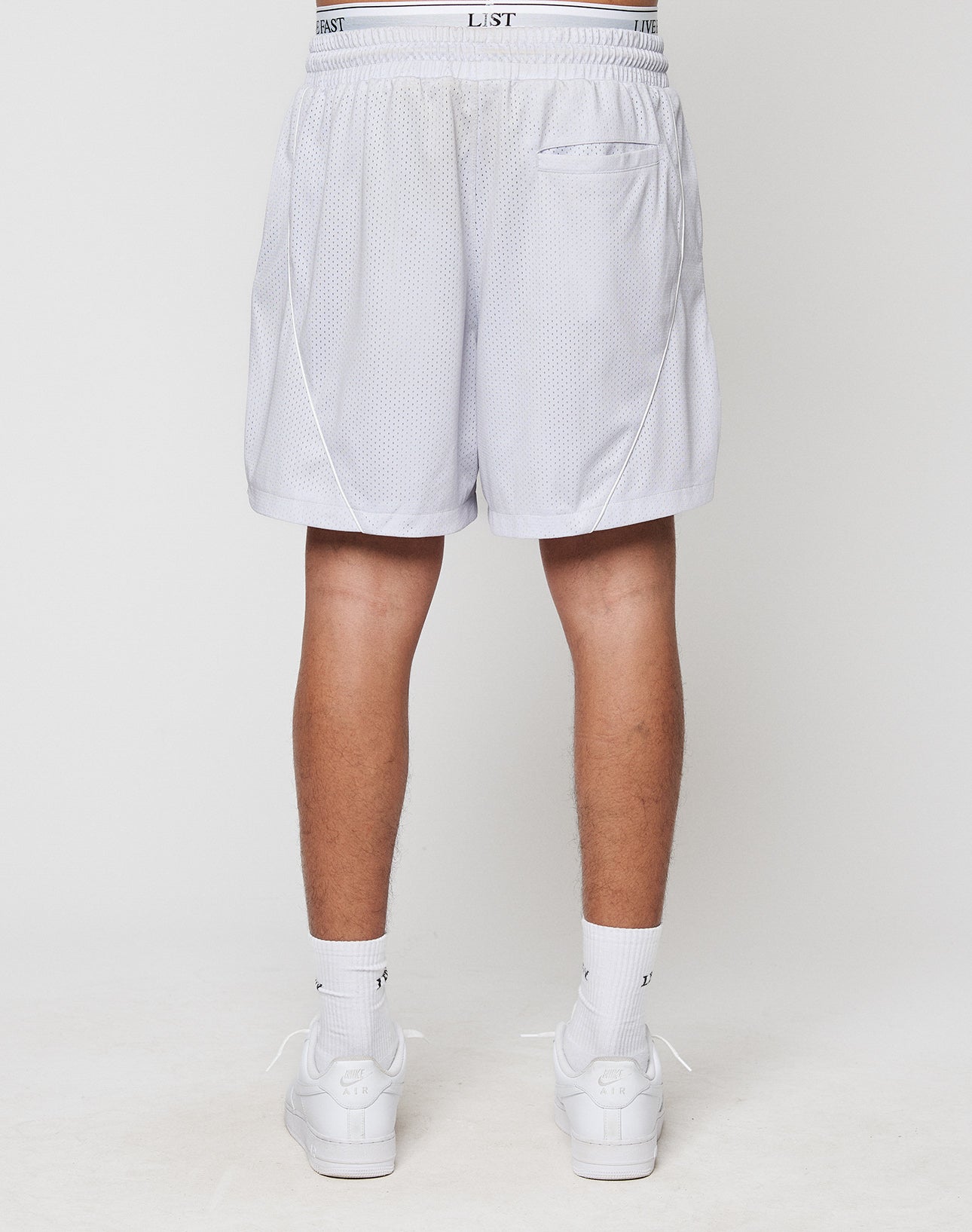 A person faces away, wearing LIVE FAST DIE YOUNG LFDY Piping Mesh Shorts in light gray with an elastic waistband, paired with white socks and sneakers. The regular fit shorts have a mesh texture and are made from polyester, set against a plain white background.